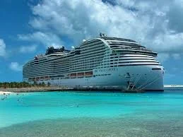 Large cruise ship docked near a tropical beach with clear turquoise water and blue sky.