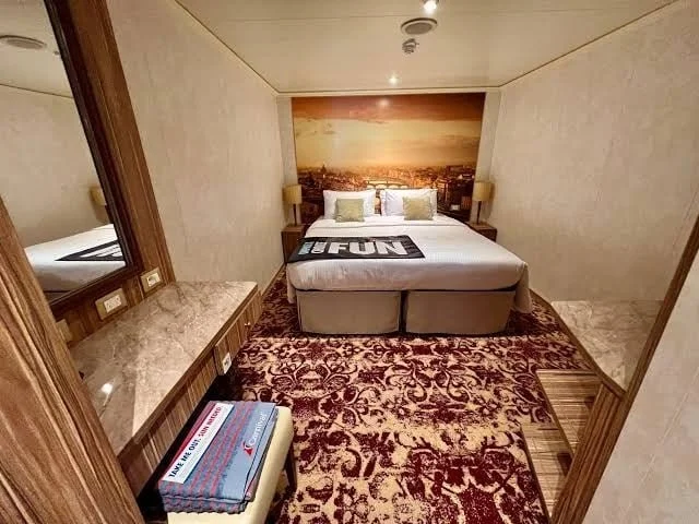 Hotel room with a bed, two nightstands with lamps, a large carpet with a floral design, a marble-topped dresser, and a mural of a cityscape behind the bed.