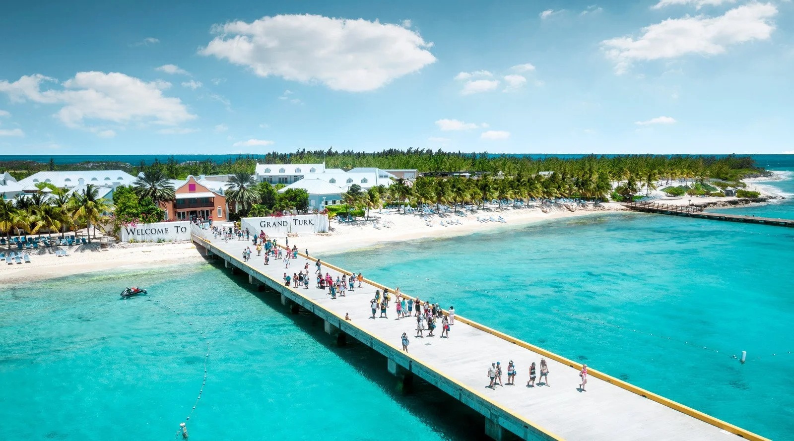 Aerial view of a tropical resort with a pier extending into clear blue water. The resort has white buildings, palm trees, and lounge chairs on the beach. A sign reads 'Welcome to Grand Turk.' People are walking along the pier, enjoying the sunny weather.