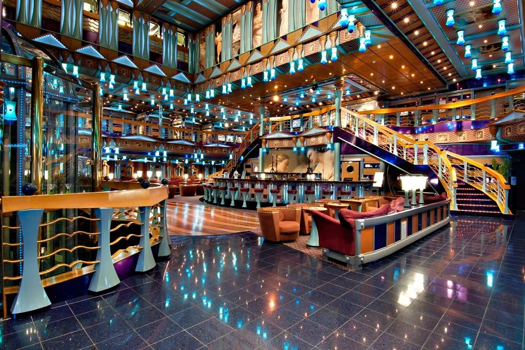 Interior of a brightly lit lounge or bar area with multiple seating areas, decorative lighting, and a stage on an upper level.