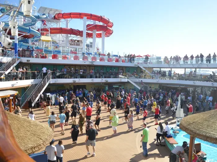 Crowd of people dancing and socializing on a cruise ship deck with water slides and pools.