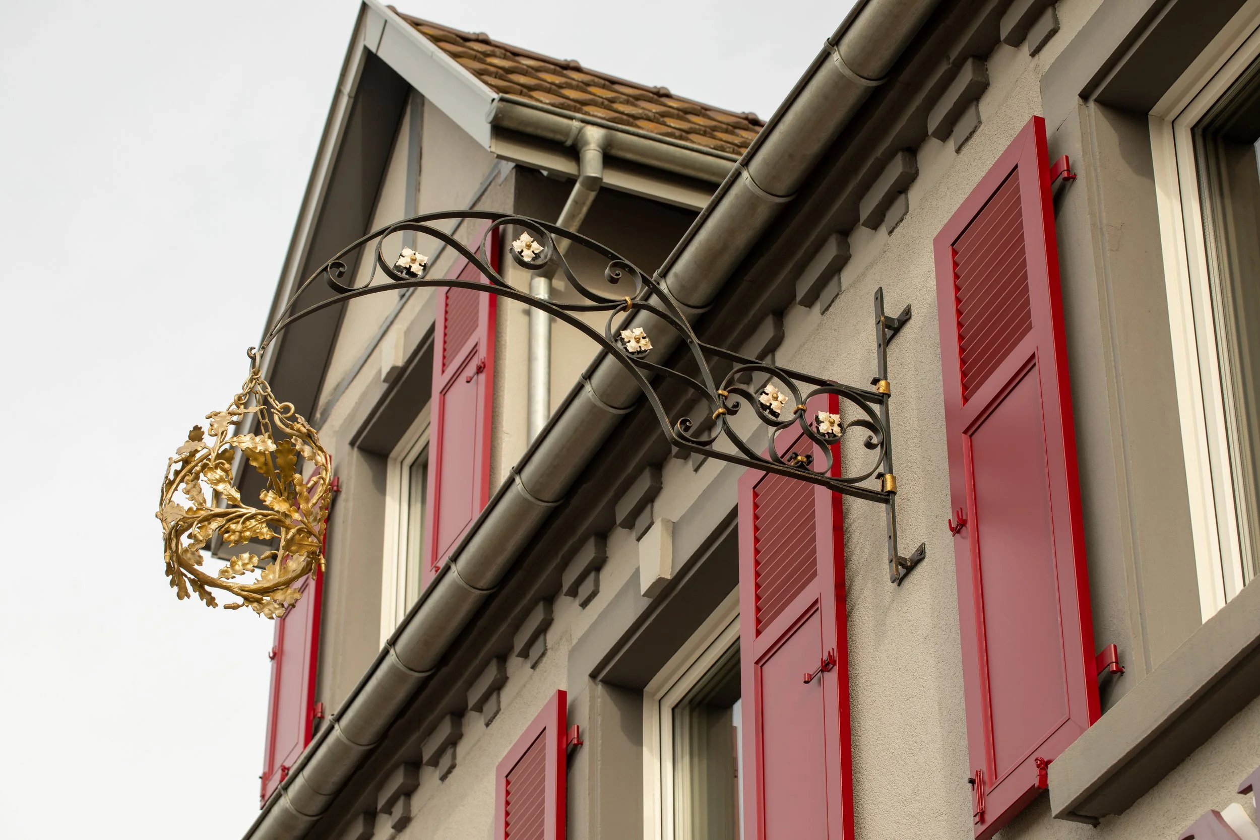 Façade du restaurant avec des volets rouges, une fenêtre, une gouttière, et une structure métallique décorative représentant une couronne suspendu 