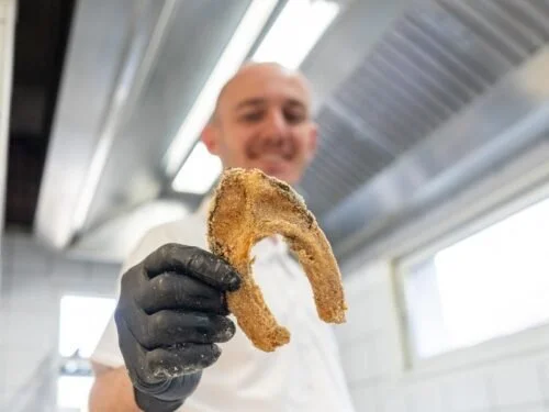 Simon Hartmann, le chef souriant tenant un morceau de friture de carpe dans un environnement de cuisine
