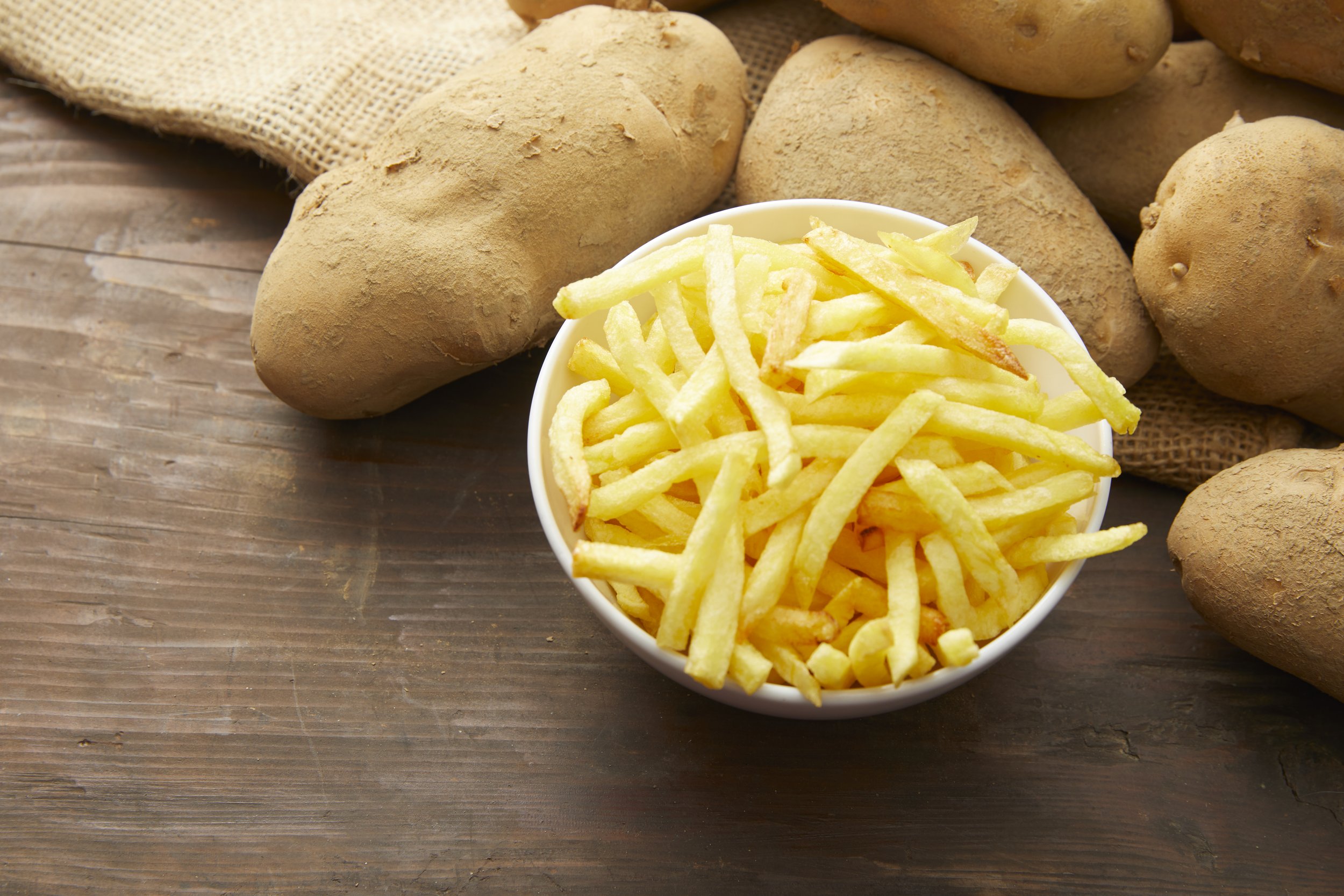Une coupe de frites dorées entourée de pommes de terre crues sur une surface en bois.