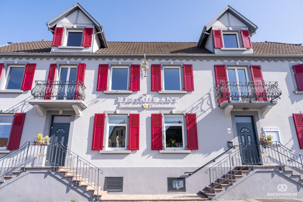 Façade du restaurant à la couronne "Hartmann Zimmermann" avec plusieurs fenêtres, volets rouges, balcons en fer forgé, deux escaliers d'accès, et une enseigne indiquant 'RESTAURANT'.