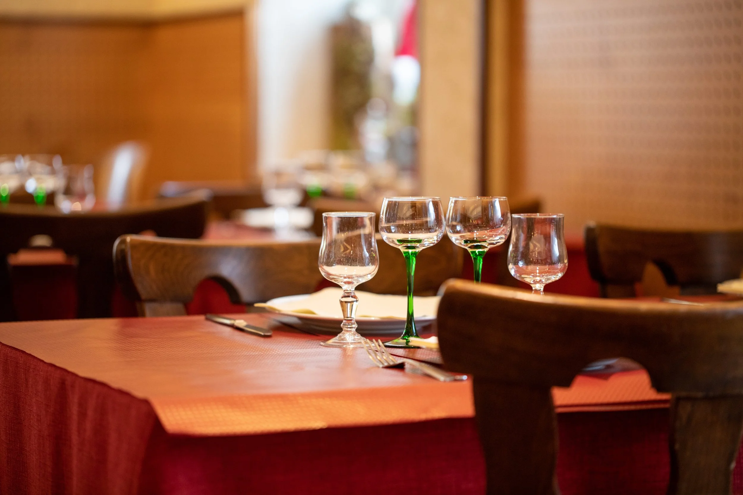 Table dressée dans un restaurant avec des verres à vin à la fois clairs et avec des tiges vertes, une fourchette et un couteau, sur une nappe orange.