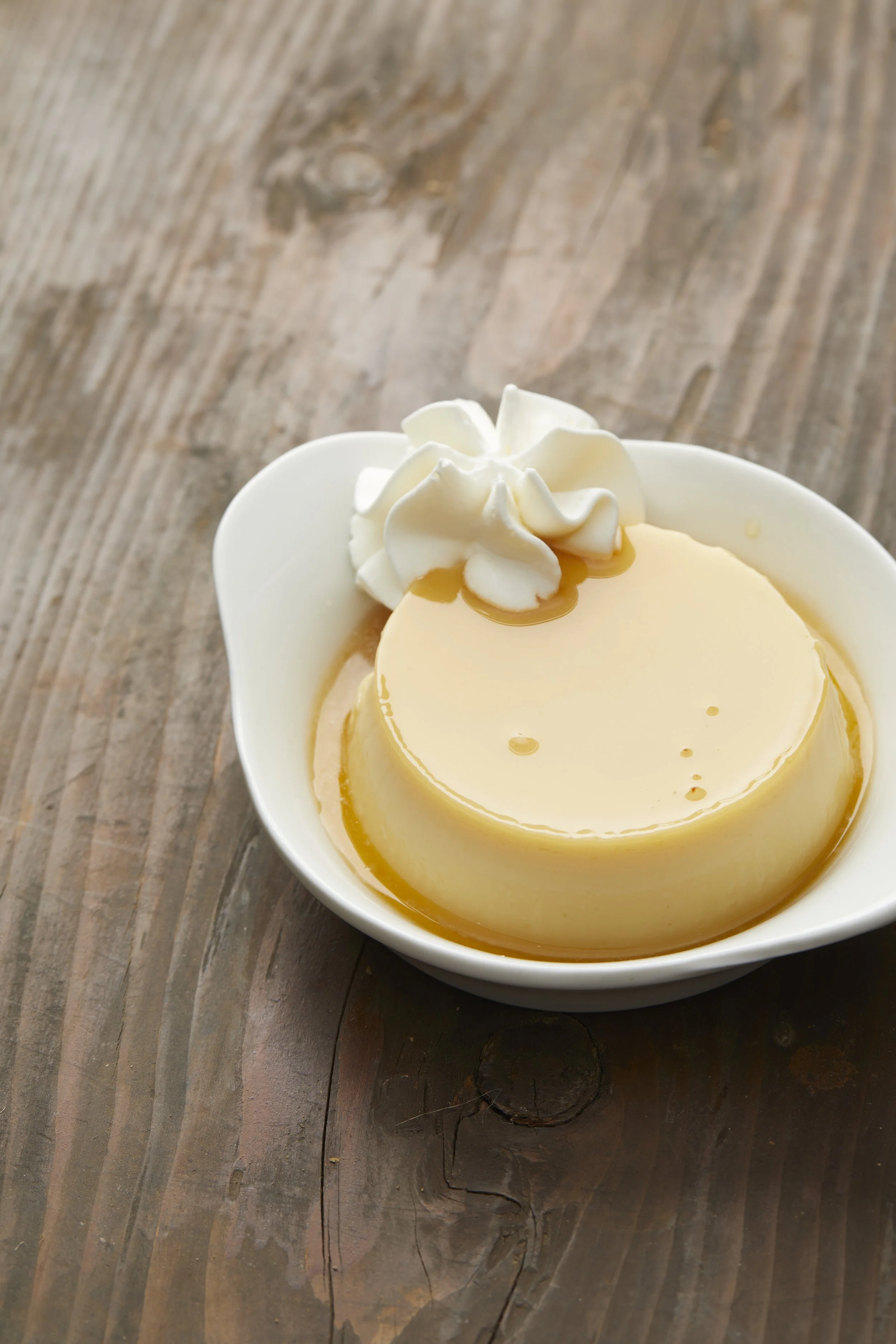 Crème caramel avec une garniture de crème chantilly et du caramel, servi dans un bol blanc sur une table en bois.