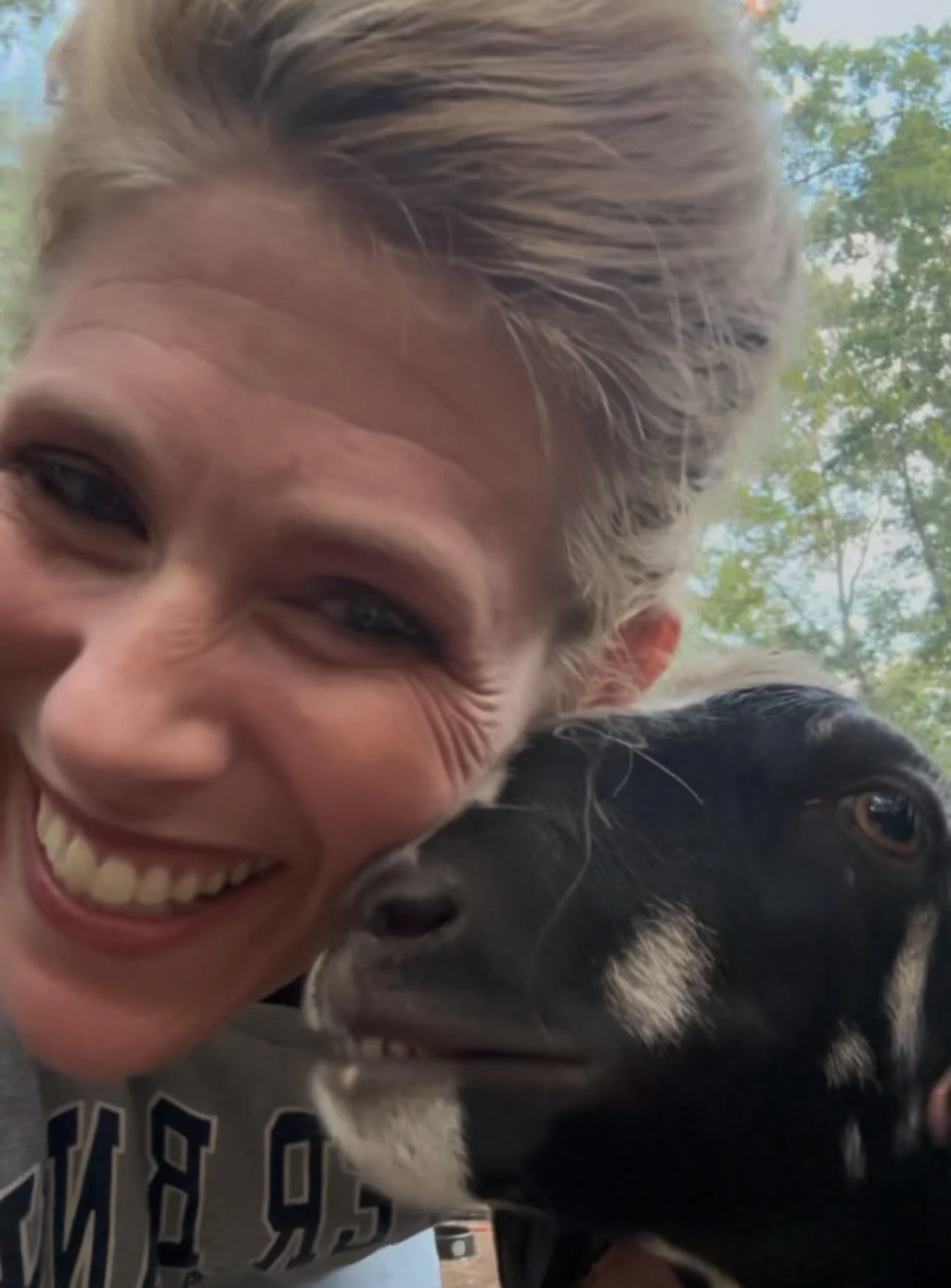 A woman smiling and hugging a black and white dog outdoors, with trees and sky visible in the background.