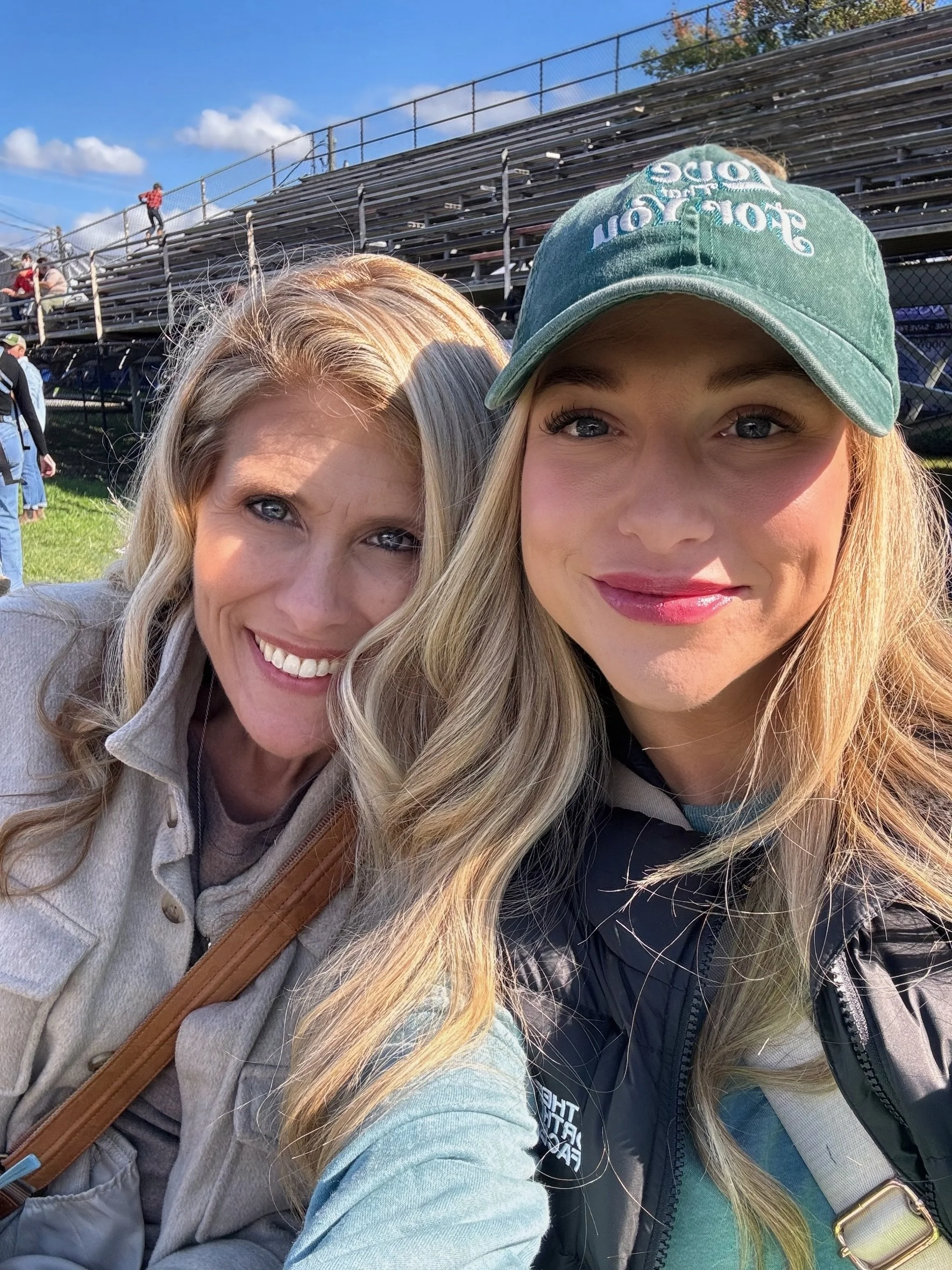 Two women taking a selfie at a sports stadium on a sunny day, with empty bleachers in the background.