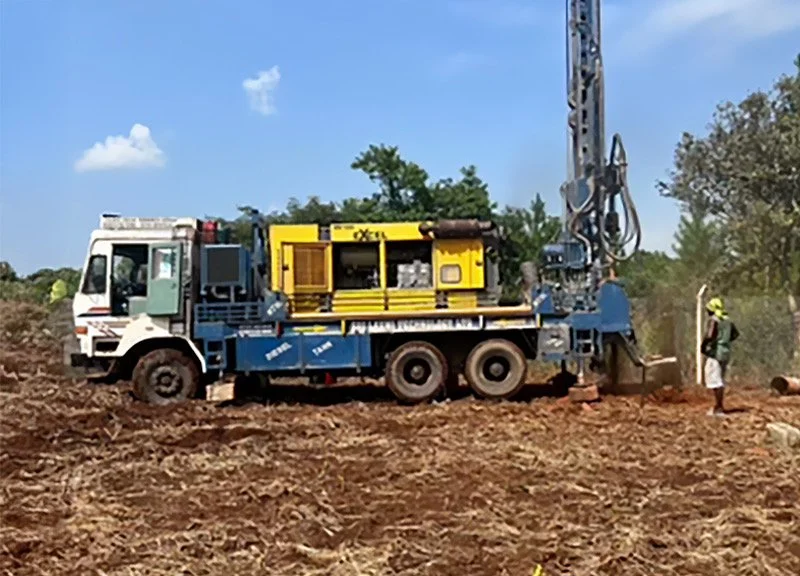 A construction site with a large drilling truck and a worker near it on dirt ground with trees and blue sky in the background.