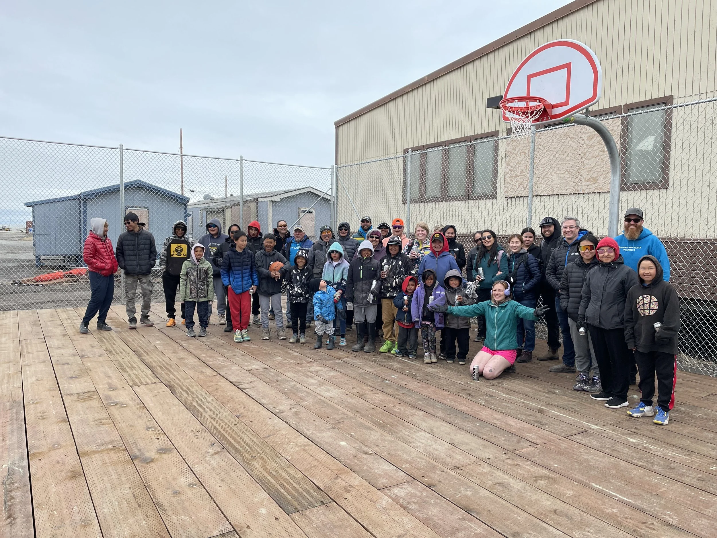 A large group of children and adults gathered outdoors on a wooden basketball court, posing for a photo in front of a chain-link fence and a basketball hoop, with cloudy skies overhead.