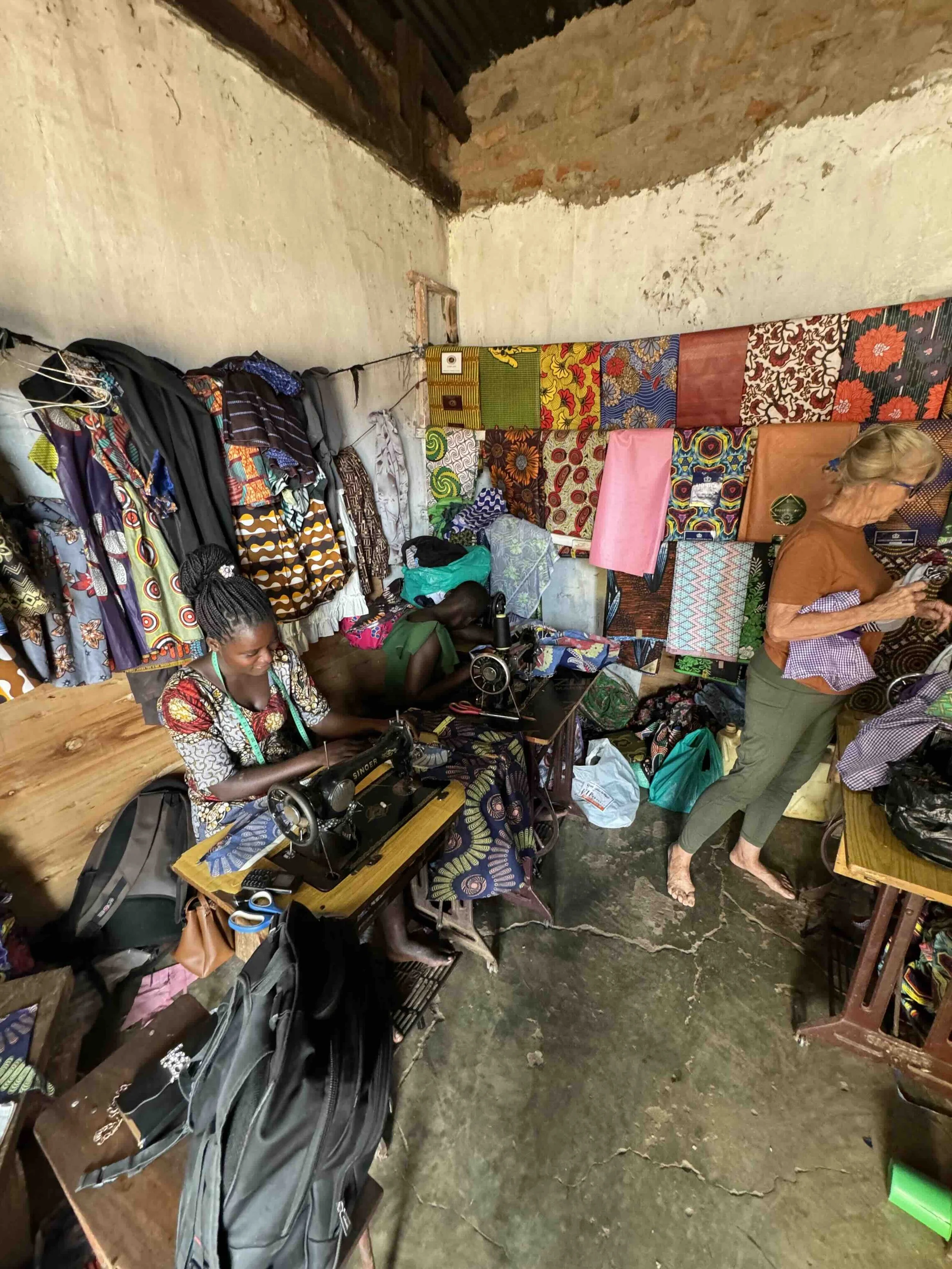 Two women working in a small sewing workshop with colorful fabric pieces hanging on the wall and sewing machines on the tables.