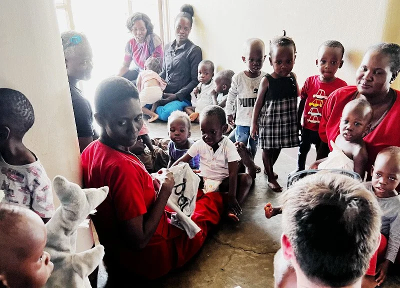 Group of children and women gathered indoors, some sitting and others standing, around a woman sewing or working with cloth.