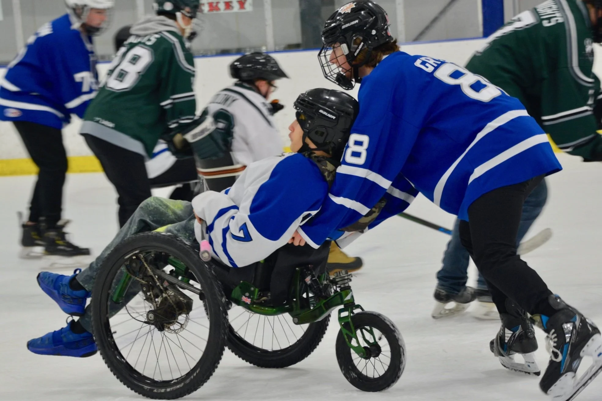 A person in a wheelchair being pushed by another person during an hockey game on ice, with other players in the background.