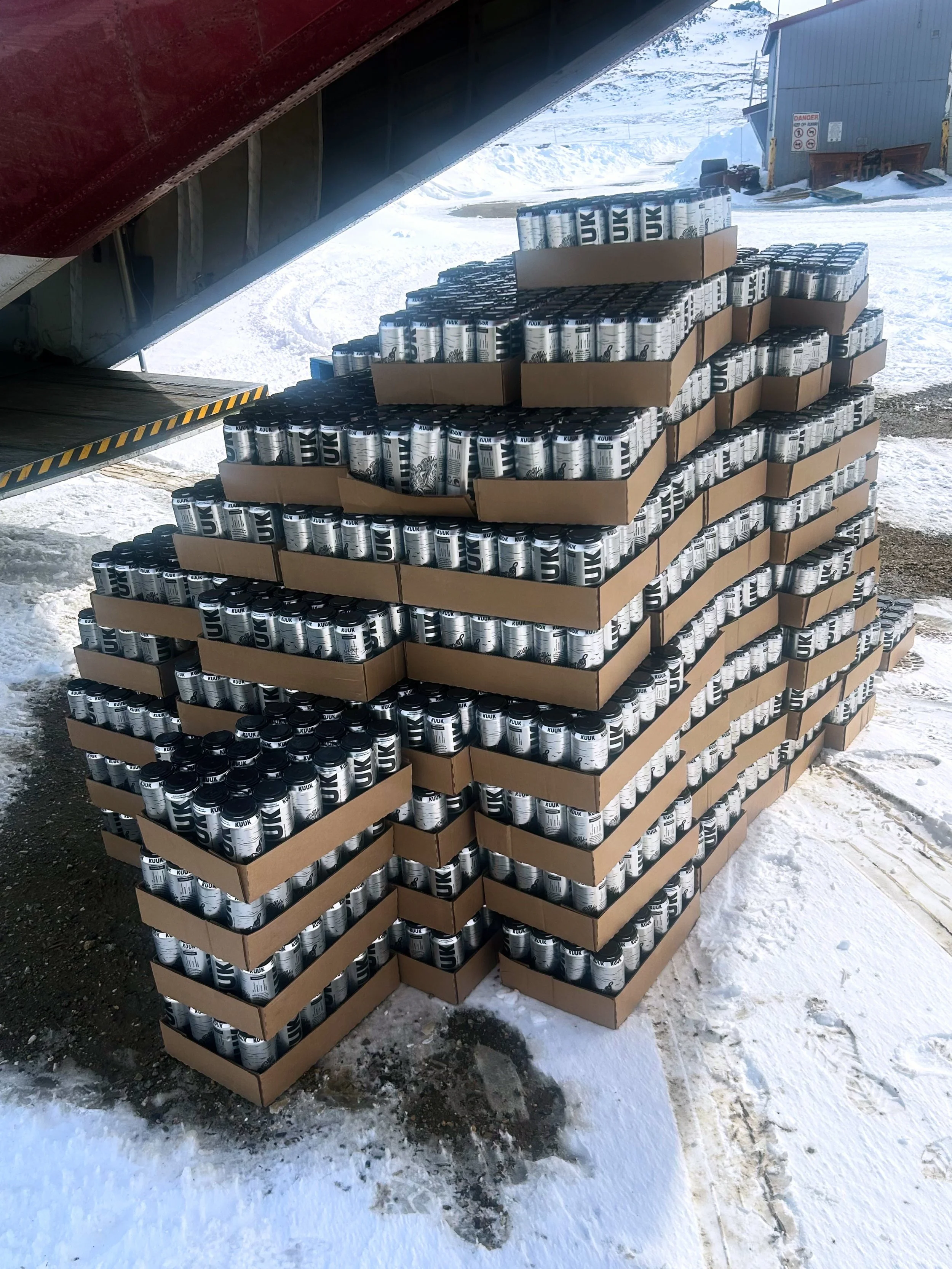Pallet of cans stacked under the rear of a plane, with a snow-covered ground outside.
