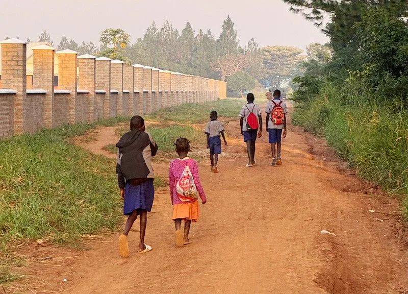 Children walking on a dirt path alongside a brick wall and green trees.
