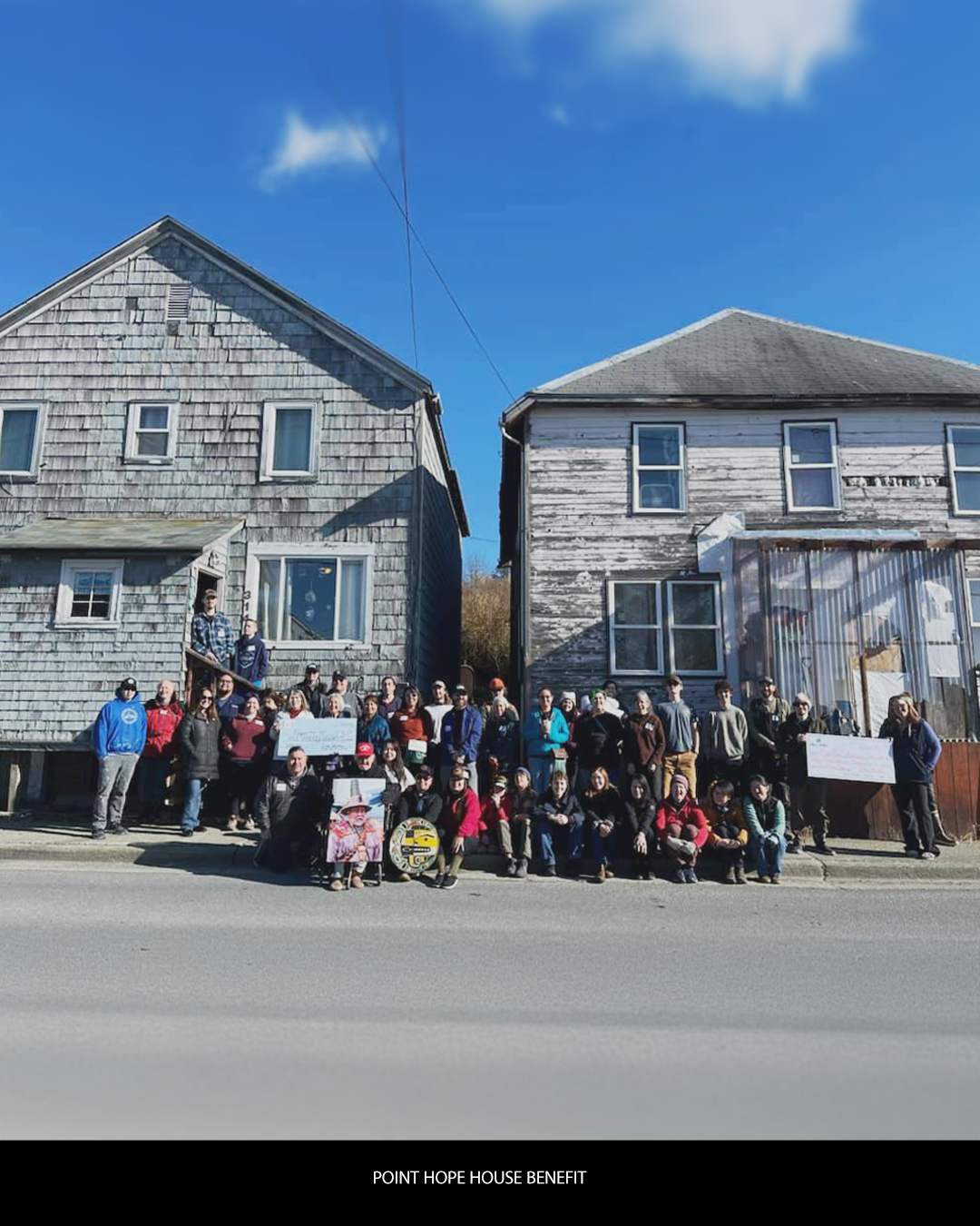 Group of people standing in front of two old wooden houses during a service event, with signs and banners.