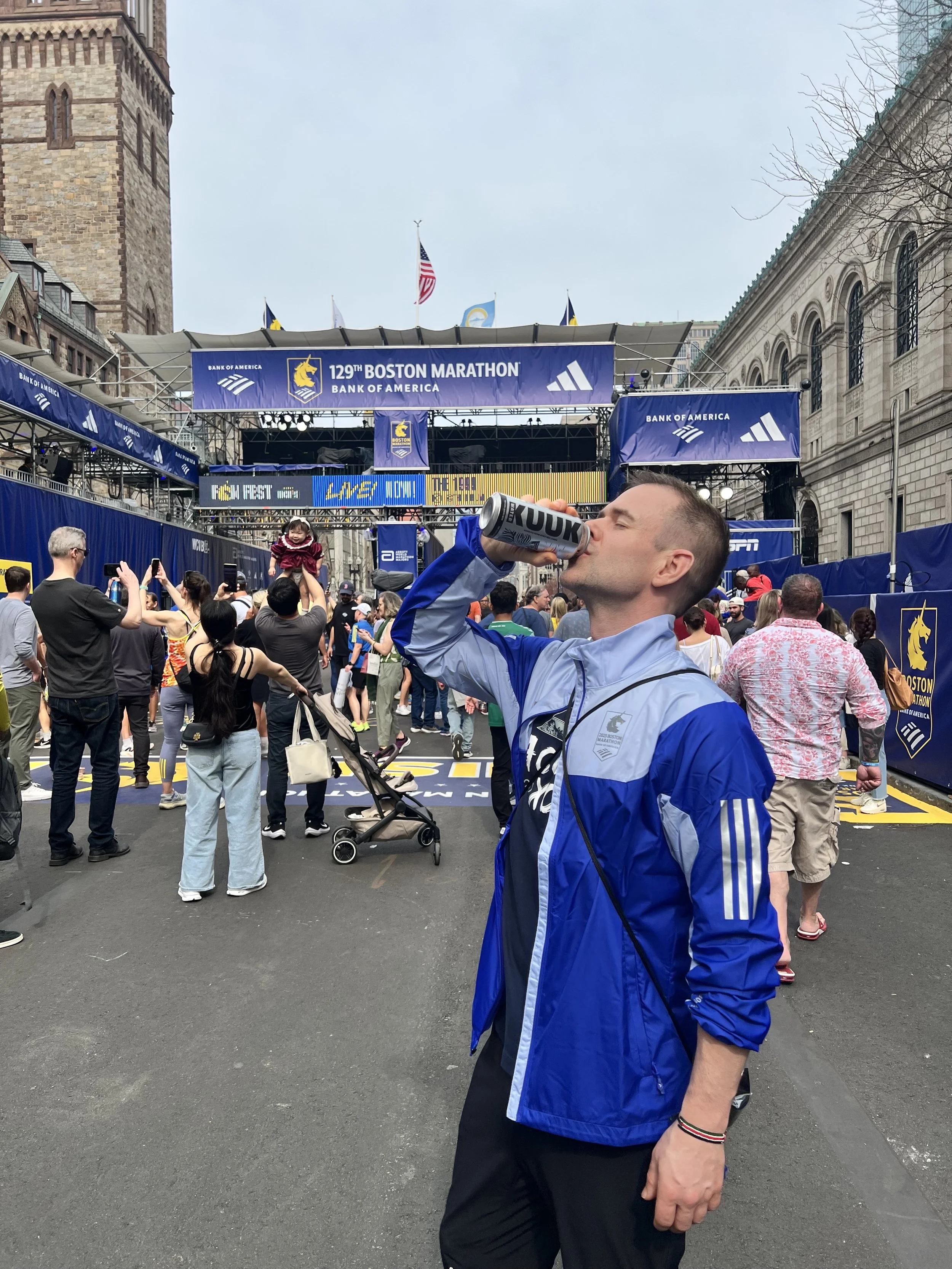 A man drinking from a can at the finish line of the 129th Boston Marathon, with a crowd of spectators and other runners around him, and a large banner overhead that reads '129th Boston Marathon'.