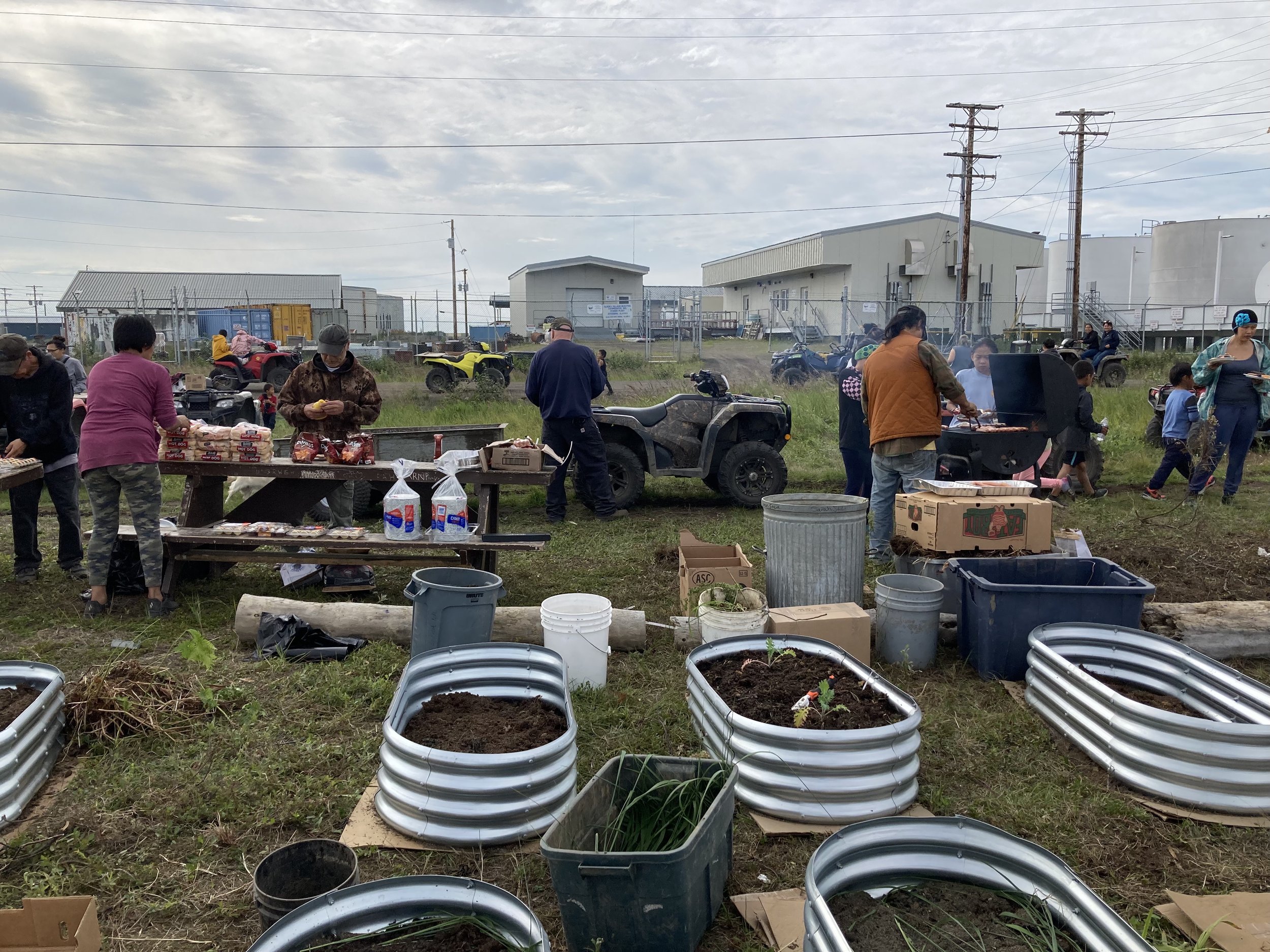 People gathered outdoors, some preparing food on grills, others sorting supplies or working with plants, in a grassy area with industrial buildings and power lines in the background.