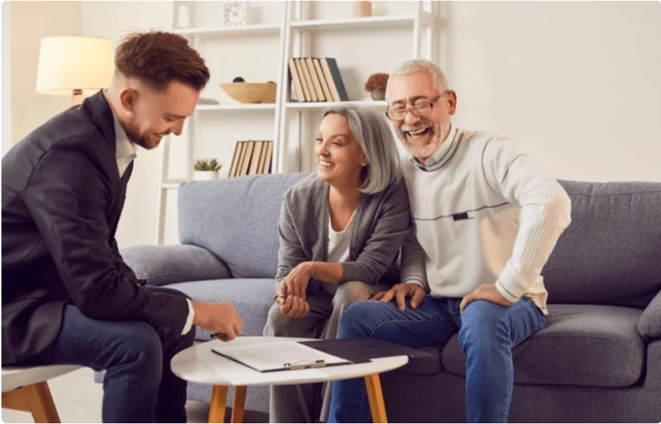 A couple sitting on a gray sofa, smiling, as a young man in a suit shows them paperwork on a small round table in a cozy living room.