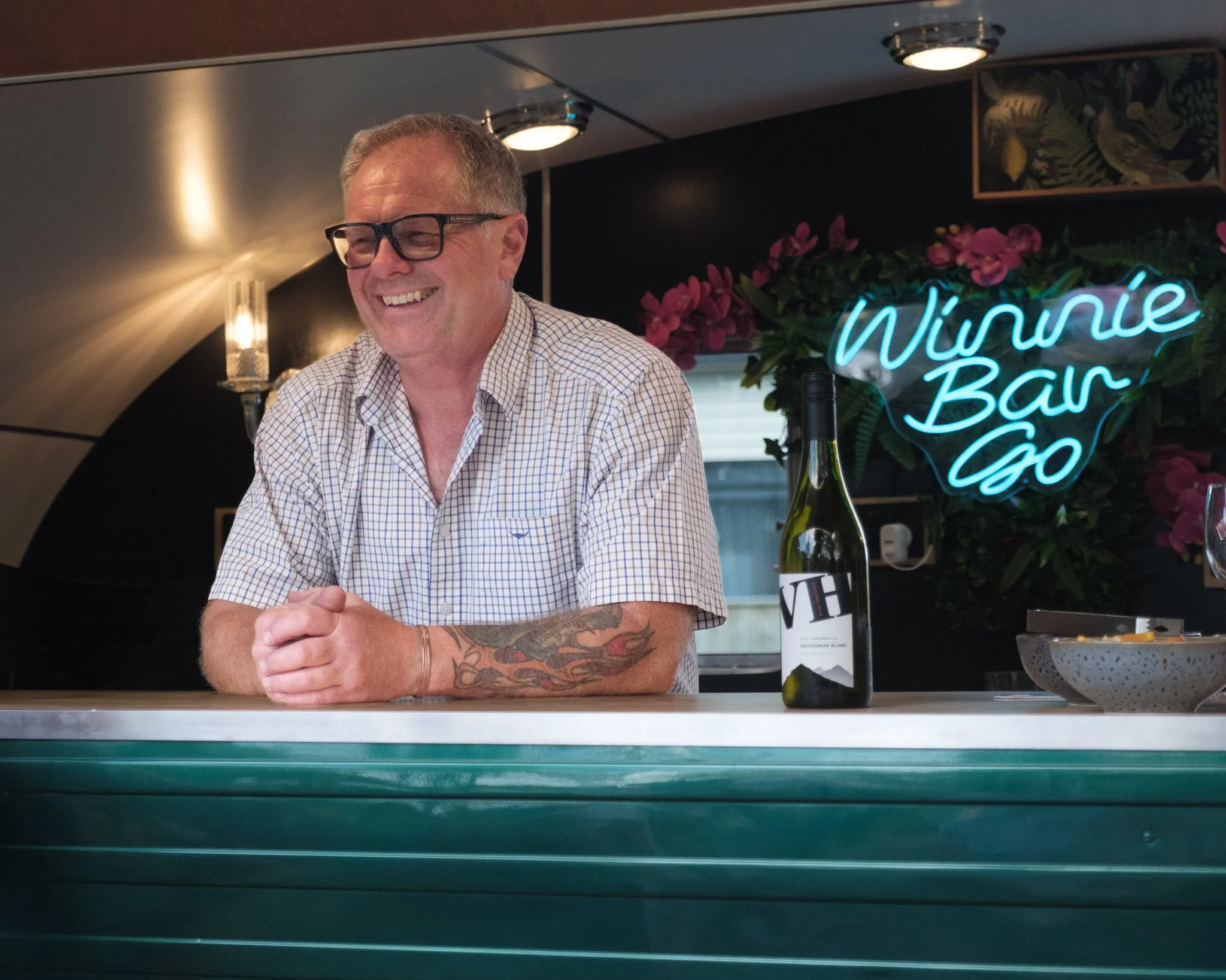 Paul standing at a bar with a bottle of wine, in front of a neon sign that reads 'Winnie-Bar-Go' with pink flowers in the background.