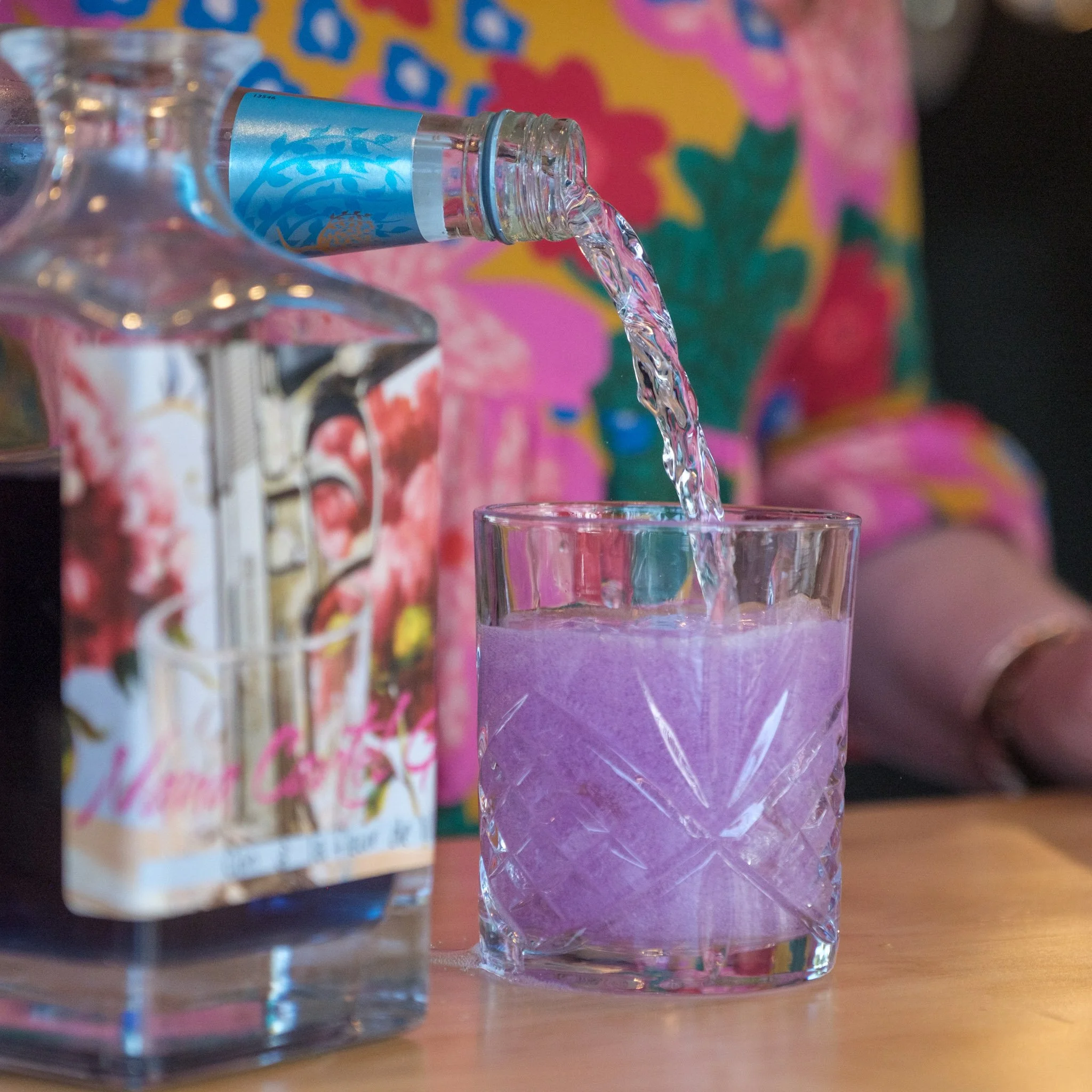 A person in a colorful floral shirt is pouring water from a decorative bottle into a purple glass on a wooden table.