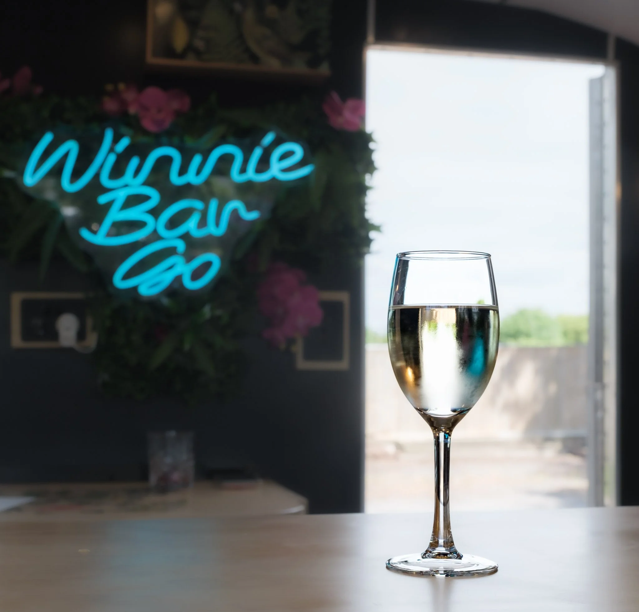 A glass of white wine on a table with a blurred background that includes a neon sign saying 'Winnie-Bar-Go'.