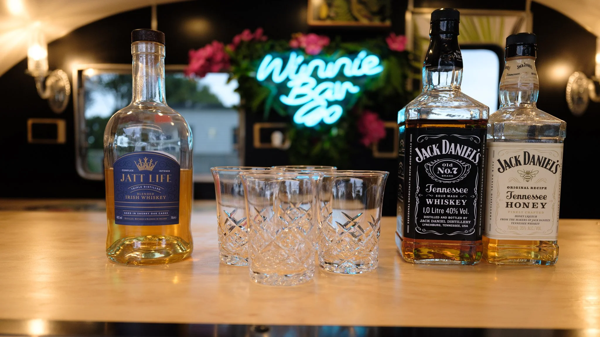A wooden table with one bottle of Irish whiskey, two bottles of Jack Daniel's whiskey, and four crystal glasses. In the background, a neon sign reads "Wrinkle Bar Co" with pink flowers above it. The setting appears to be a bar or lounge.