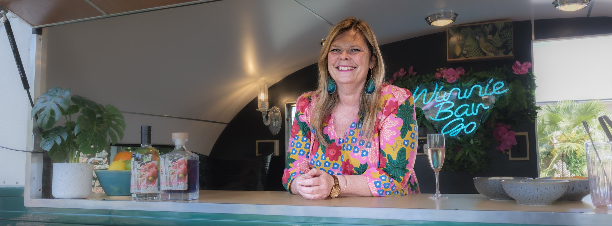 Angela smiling behind a bar counter, decorated with a large green plant, bottled drinks, a bowl of fruit, and a glass of champagne; there's a neon sign that says 'Winnie-Bar-Go' and orbs of light on the ceiling.