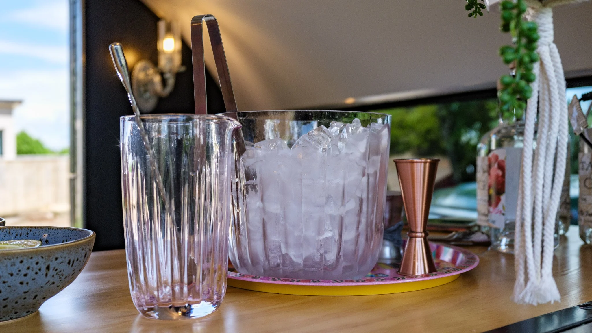 A glass container filled with ice on a countertop, with a copper jigger nearby. Other glassware and decorative items are also on the counter.