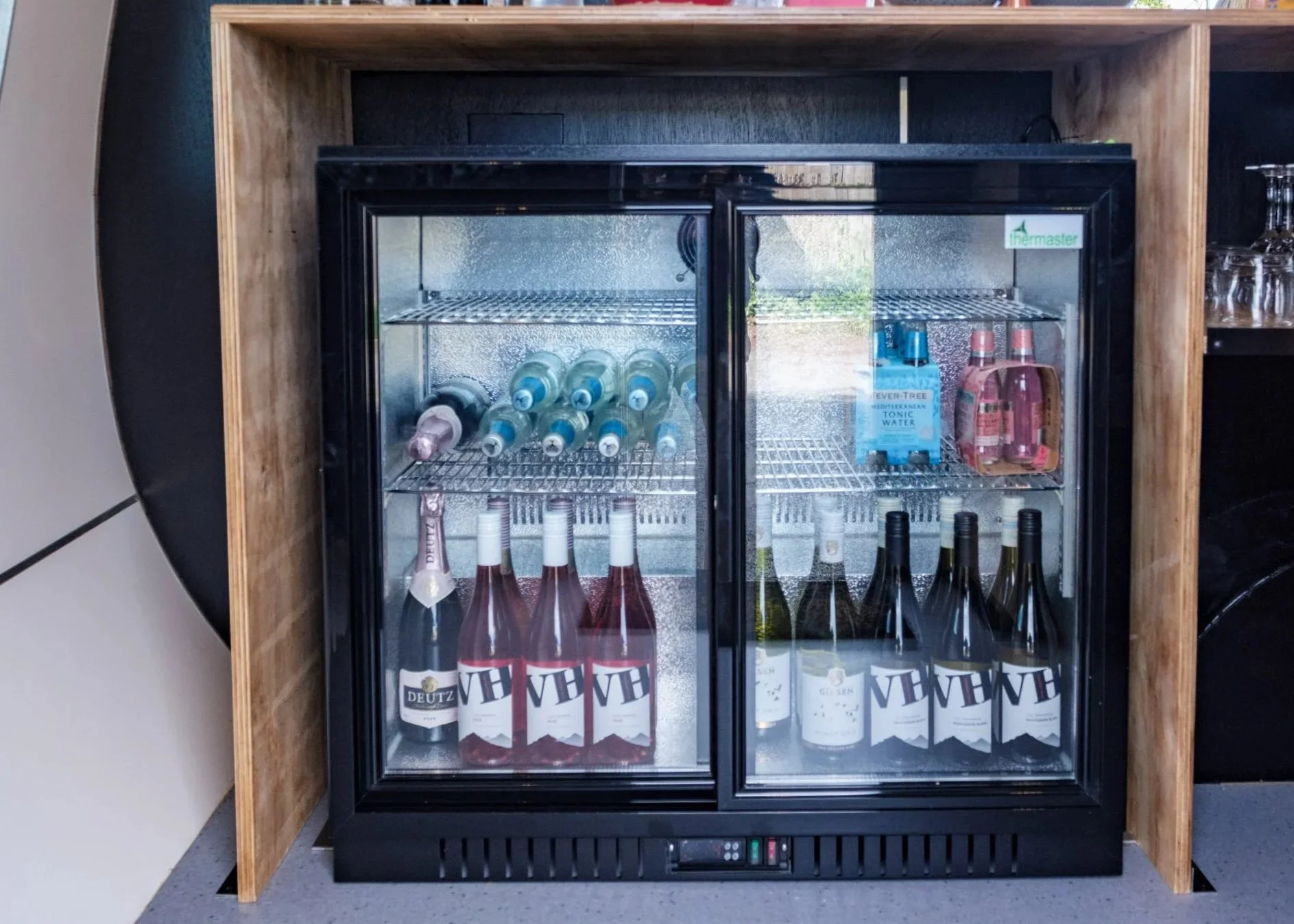 A black glass-front mini refrigerator containing bottled beverages, placed against a wooden backdrop in a setting that appears to be a bar or kitchen area.