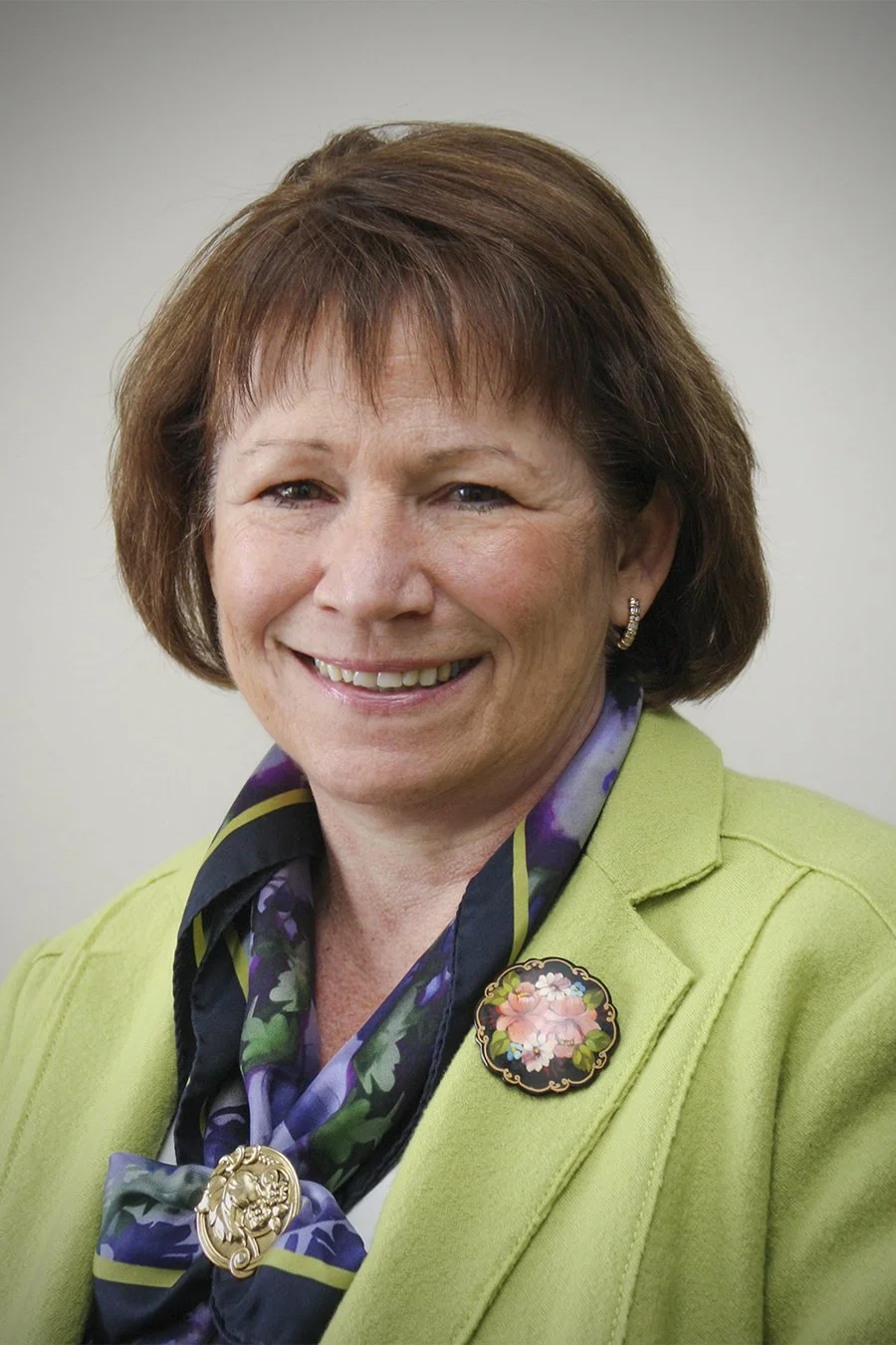 A woman with short brown hair smiling, wearing a green blazer and a purple floral scarf, with a decorative brooch.