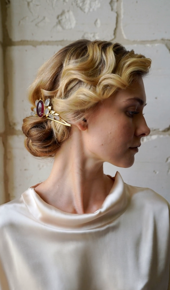 A woman with blonde, vintage-style curled hair adorned with an ornate hairpin, wearing a silky white top, standing against a white brick wall.