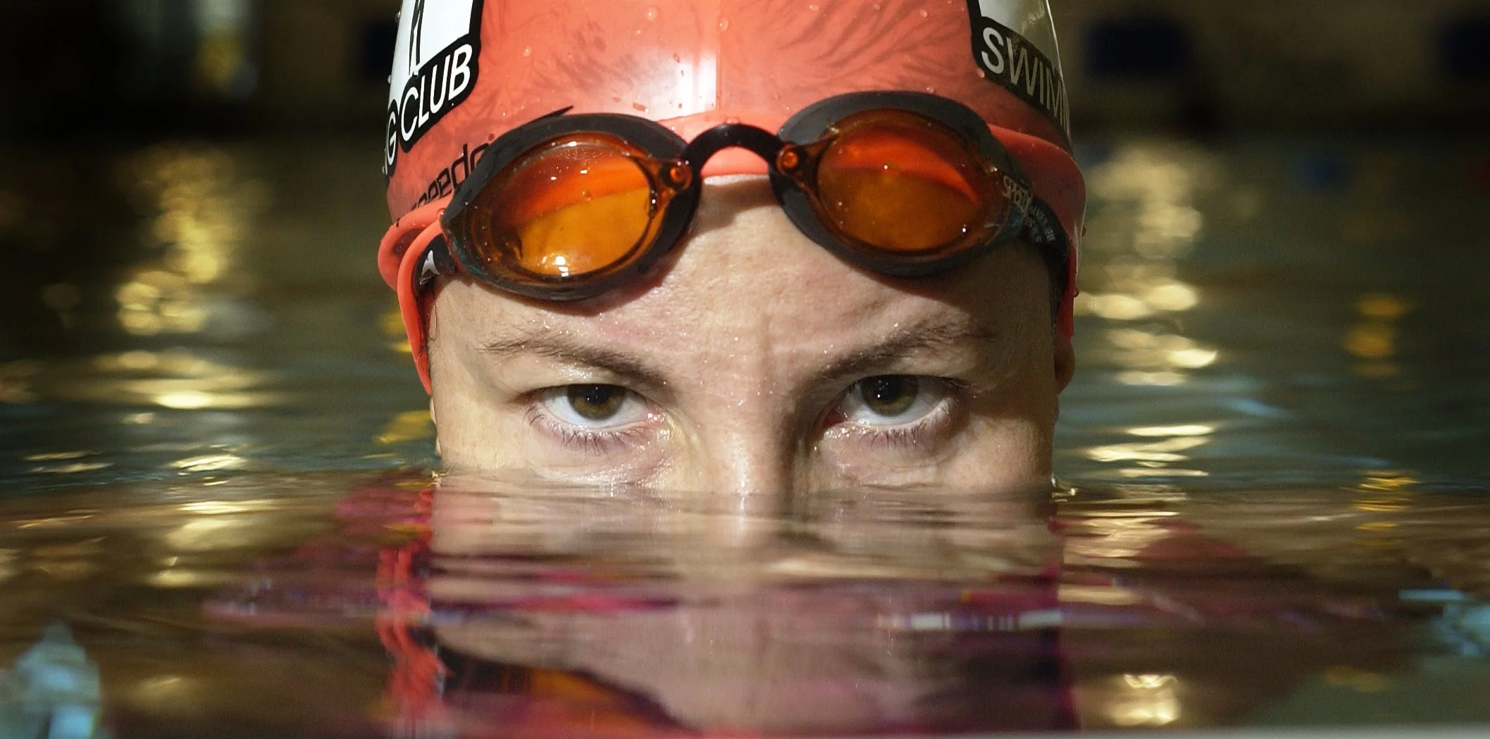 Close-up of a swimmer's face submerged in water, wearing orange goggles and a red swim cap with black and white text.