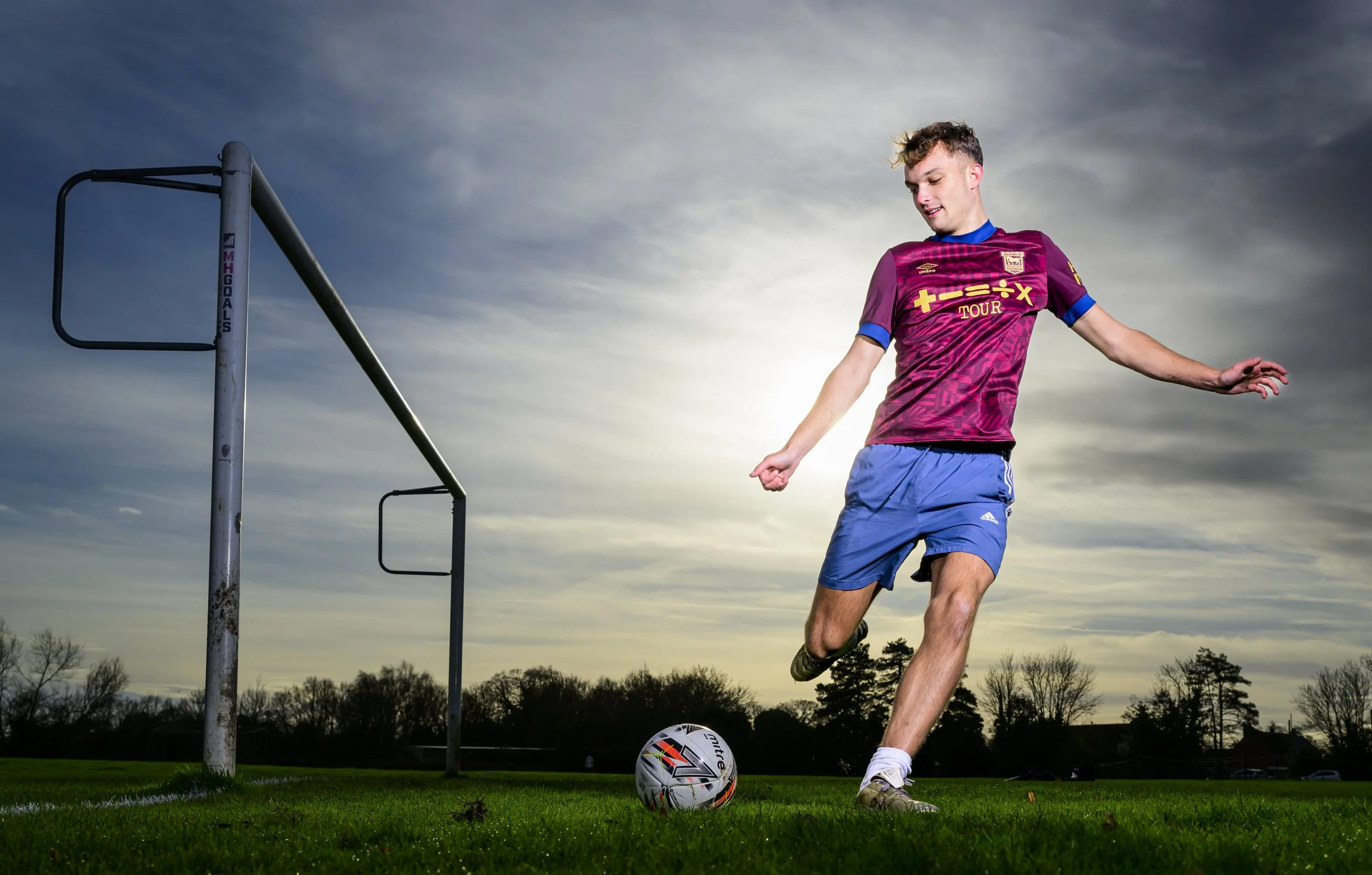A young man in a maroon and blue soccer uniform is kicking a soccer ball on a grassy field during sunset.