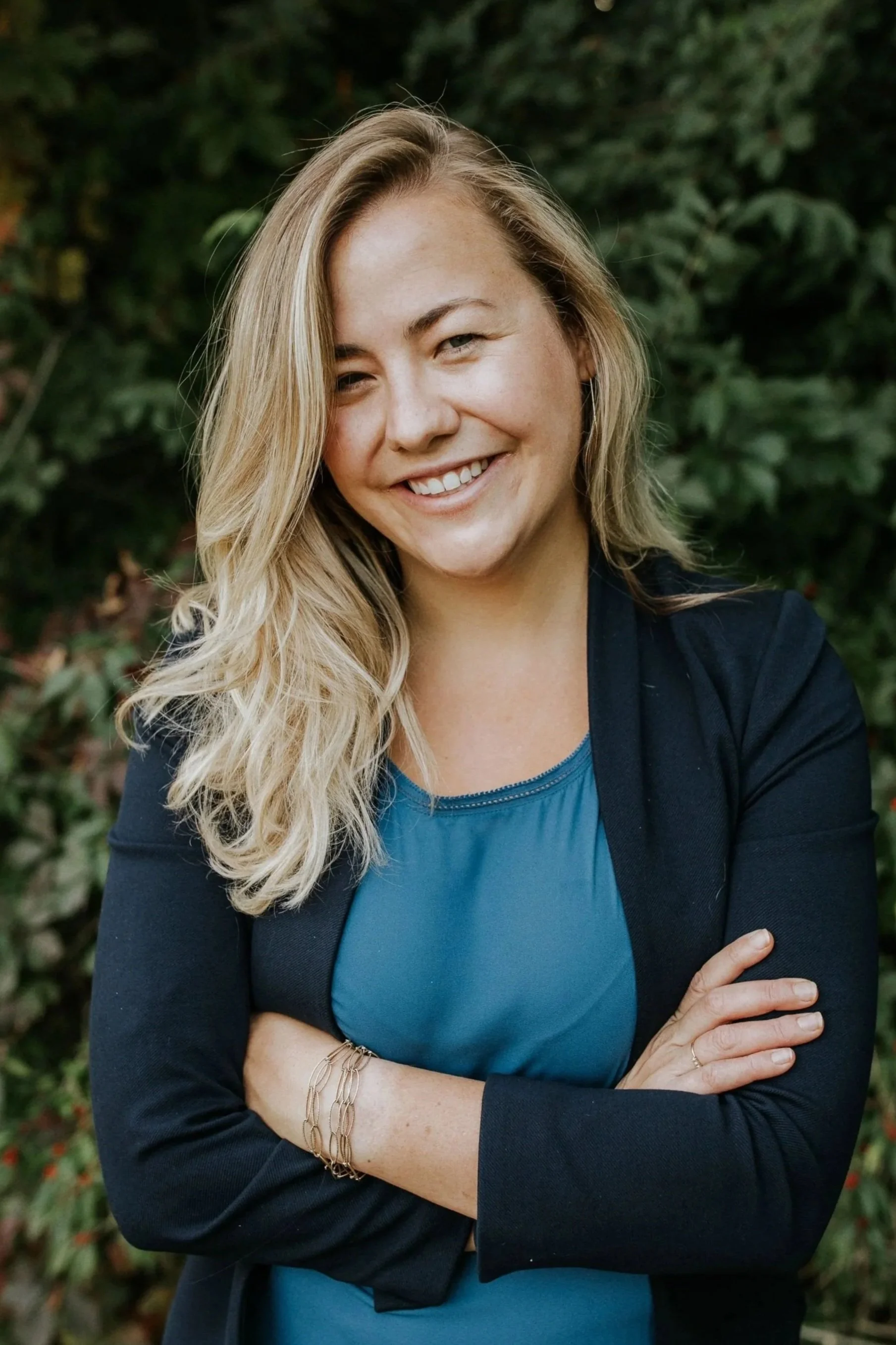 A woman with blonde wavy hair smiling with her arms crossed, wearing a black blazer and a blue top, standing outdoors with green foliage in the background.