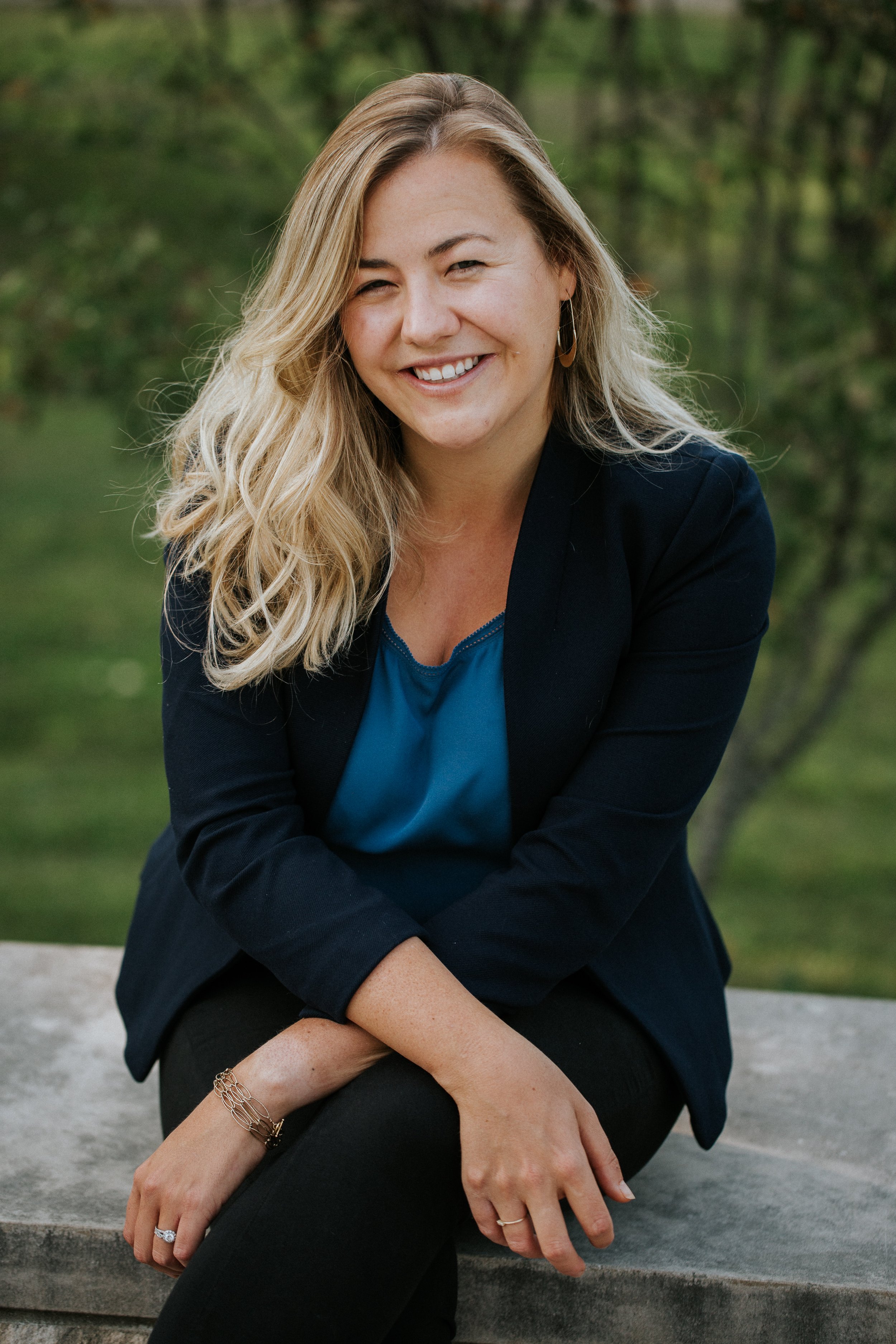 A woman with long, wavy blonde hair sitting outdoors on a stone surface, smiling at the camera. She is wearing a black blazer, a teal blouse, black pants, and jewelry, with a green background.