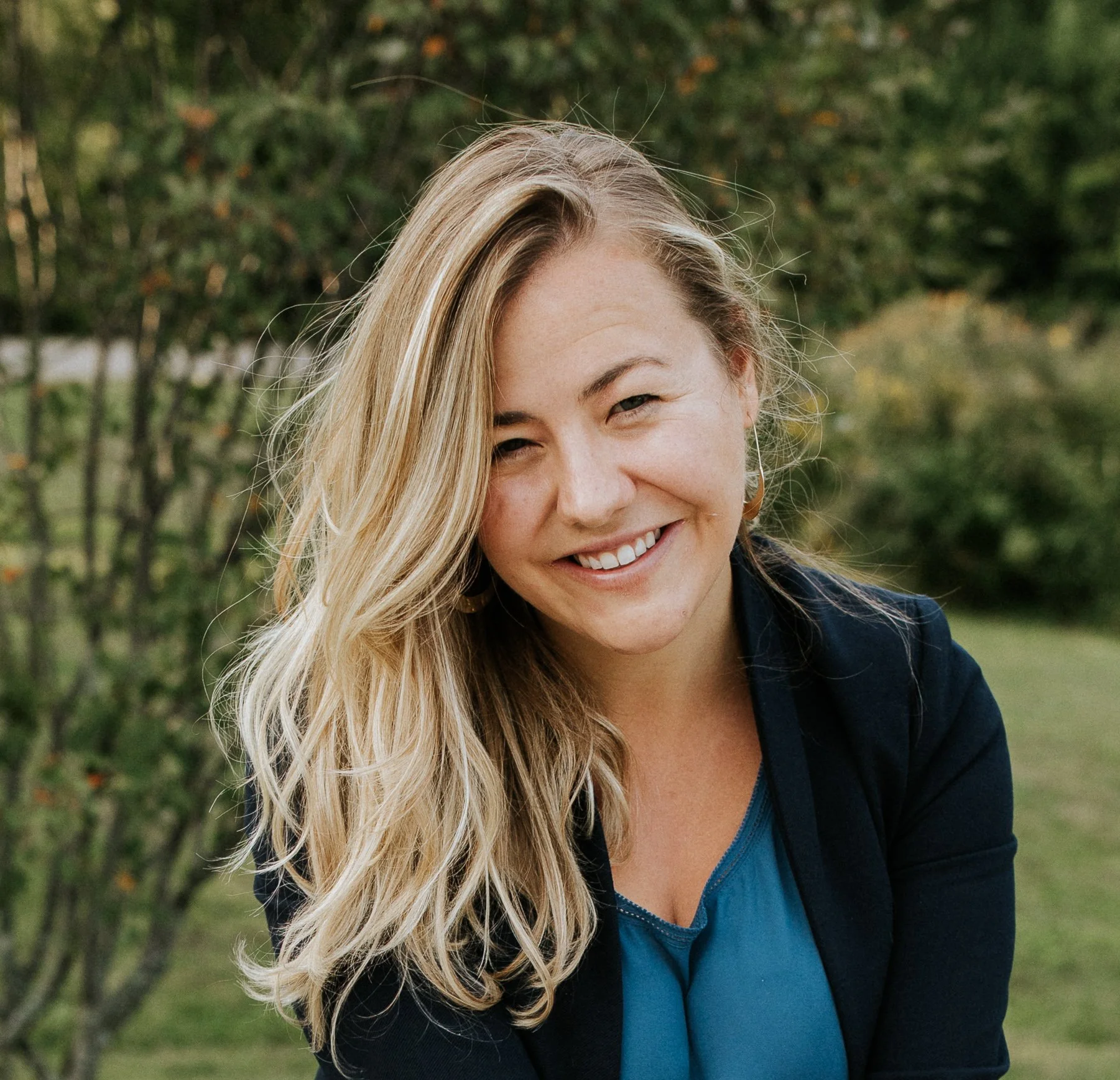 A smiling compassionate woman with long blonde hair outdoors in front of trees, wearing a black blazer and blue top.