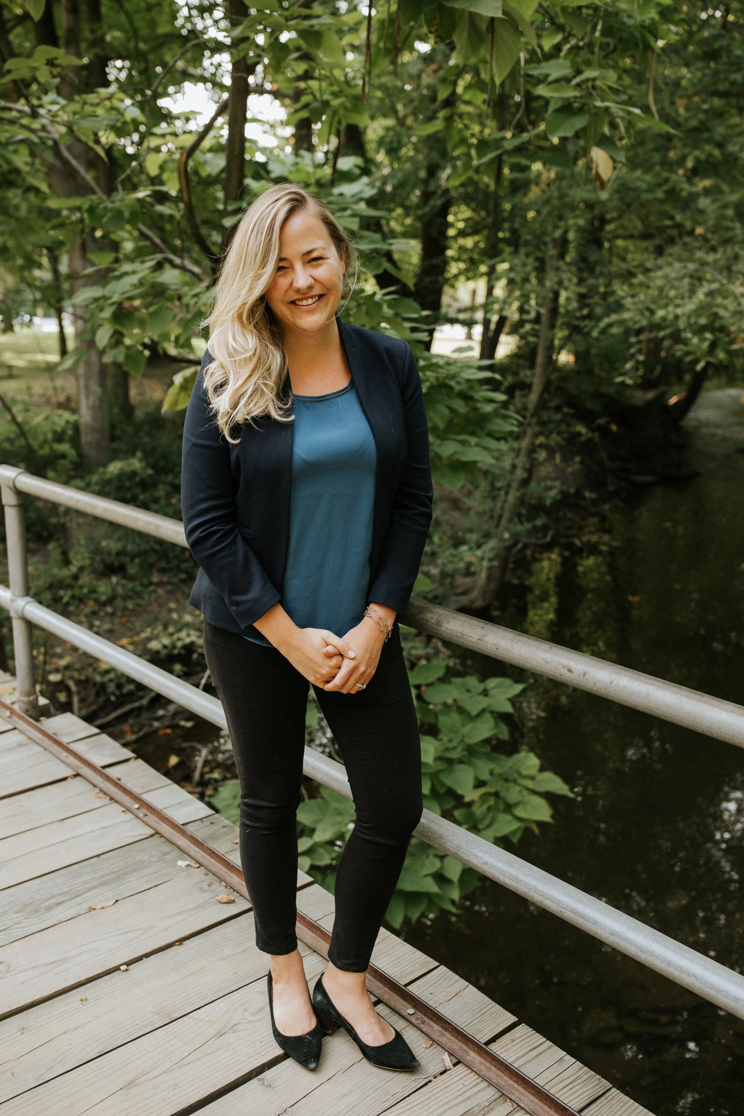 A woman with blonde hair smiling, wearing a dark blazer and teal top, standing on a wooden bridge next to a body of water surrounded by green trees.