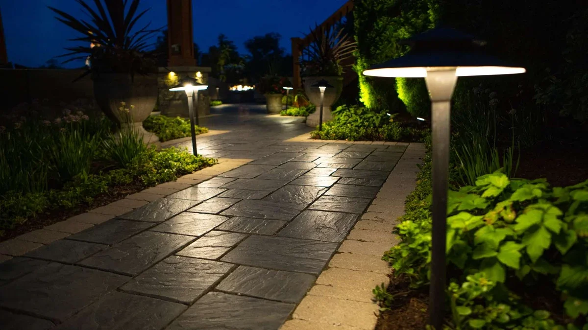 Nighttime view of a paved garden pathway lit by low garden lights, with greenery and potted plants along the sides.