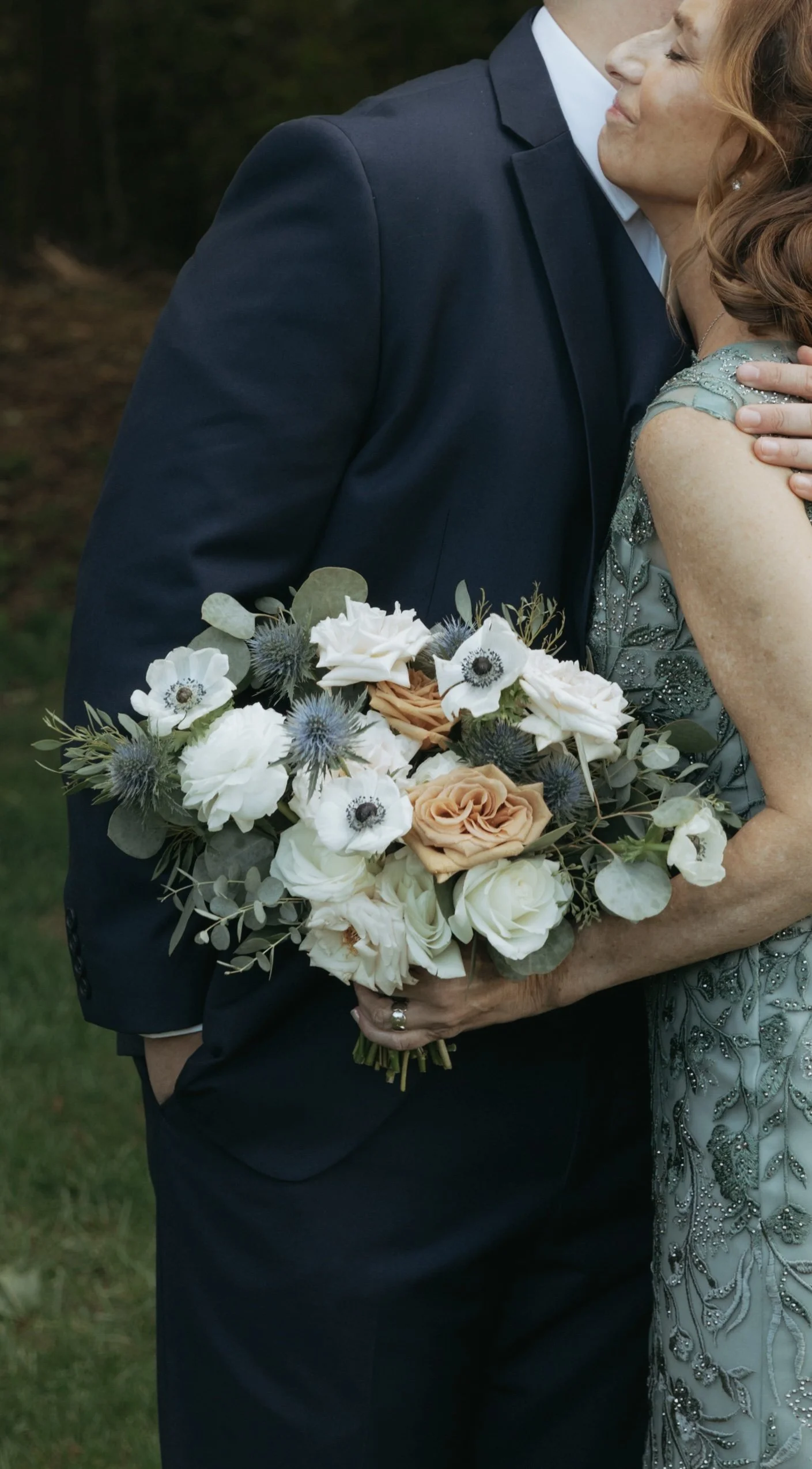 Classic white and blush bridal bouquet with roses and eucalyptus shown against a couple embracing while wearing a dark suit and green dress