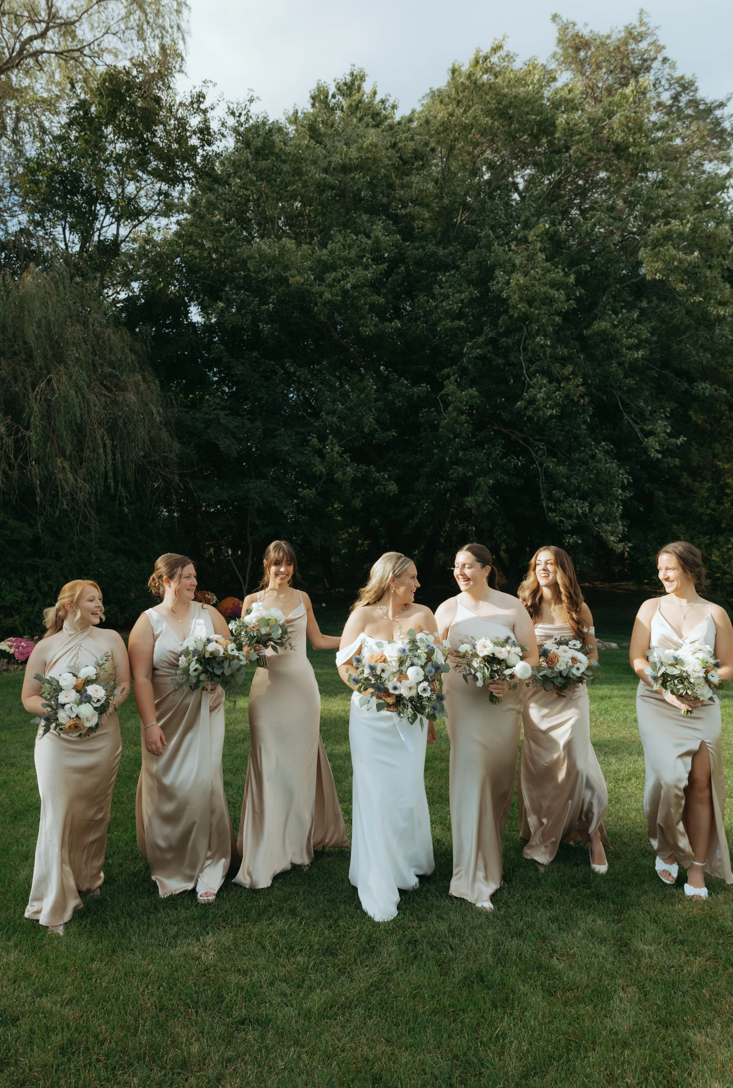 Bridesmaids in champagne gowns holding white and green bouquets outdoors.