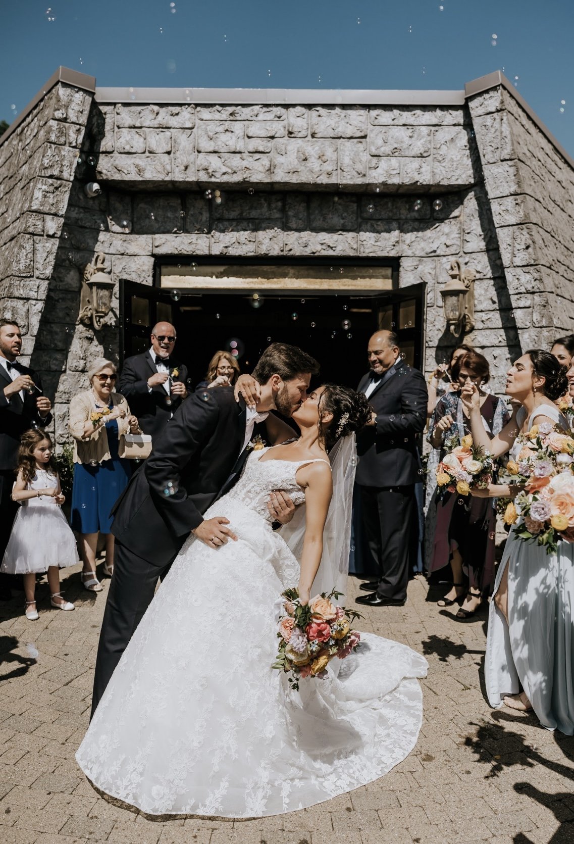 Wedding ceremony exit with confetti and guests celebrating newlyweds kissing while bride holds a gorgeous bouquet 