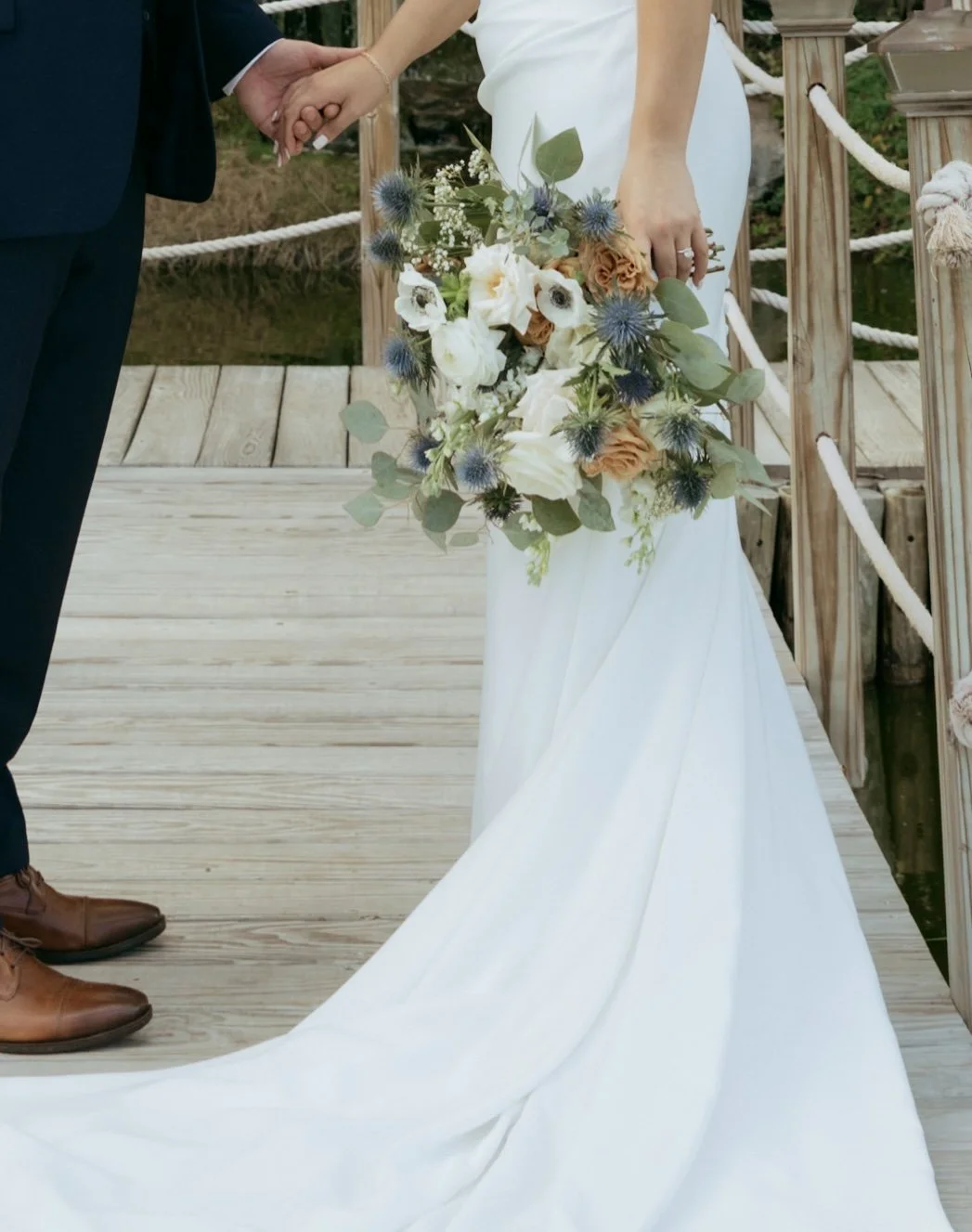 Bride with long train holding white, blush and greenery bouquet on wooden dock.