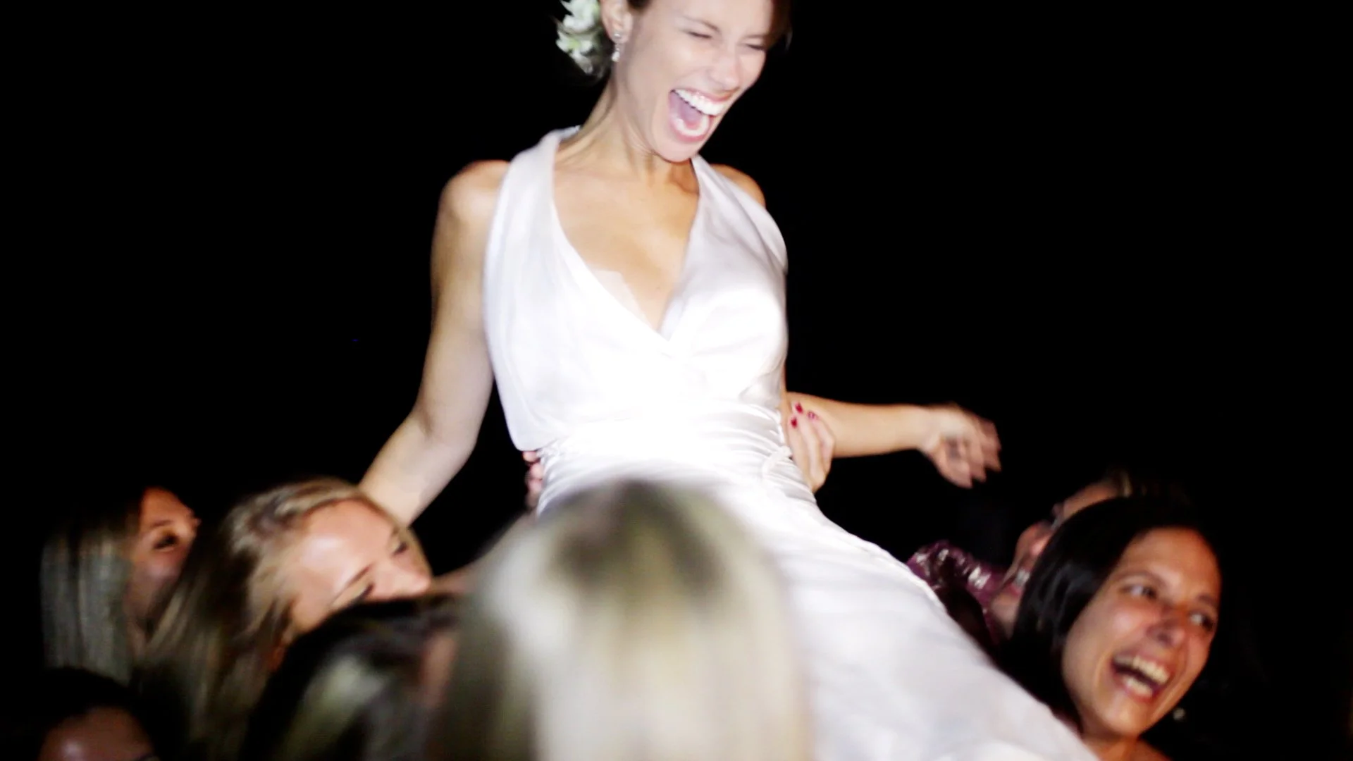 Pessoa em vestuário branco sendo levantada por um grupo de pessoas, todas sorrindo e aparentando felicidade, contra fundo preto.