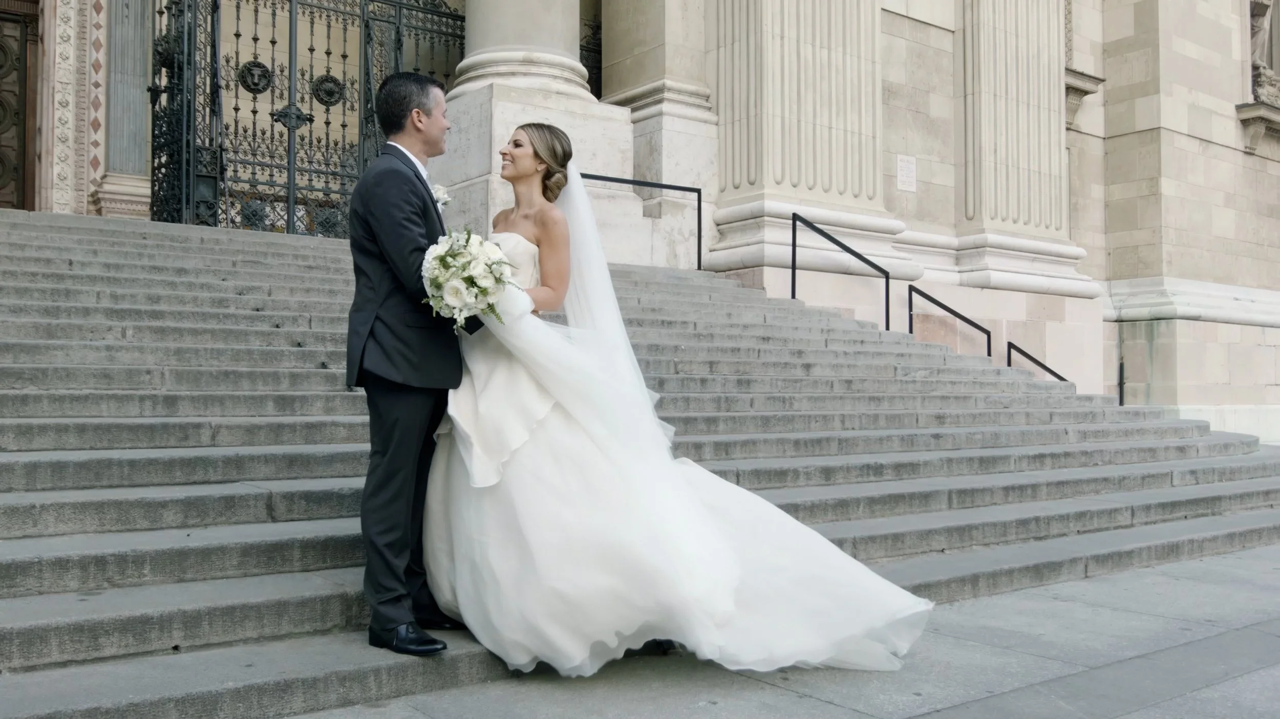 Casal de noivos sorrindo na igreja antiga, a noiva com vestido branco longo e véu, segurando um buquê de flores brancas, na escadaria de pedra da igreja com pilares e detalhes arquitetônicos clássicos.