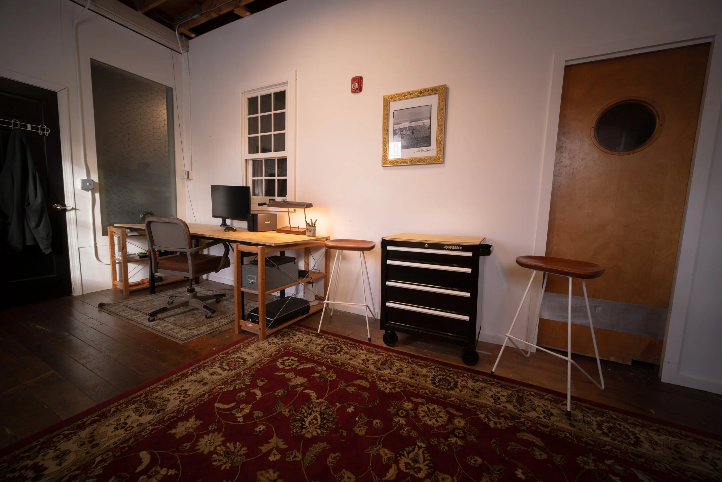 Home office with a wooden desk holding a computer monitor, keyboard, and desk lamp, two stools, a rolling black tool chest, and a patterned red area rug.