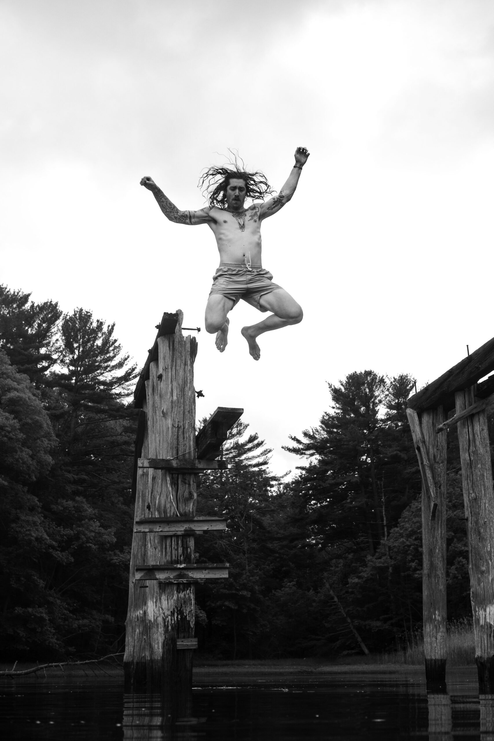 A shirtless man with long hair and tattoos jumps off an old wooden dock post into a body of water, with trees and a cloudy sky in the background.