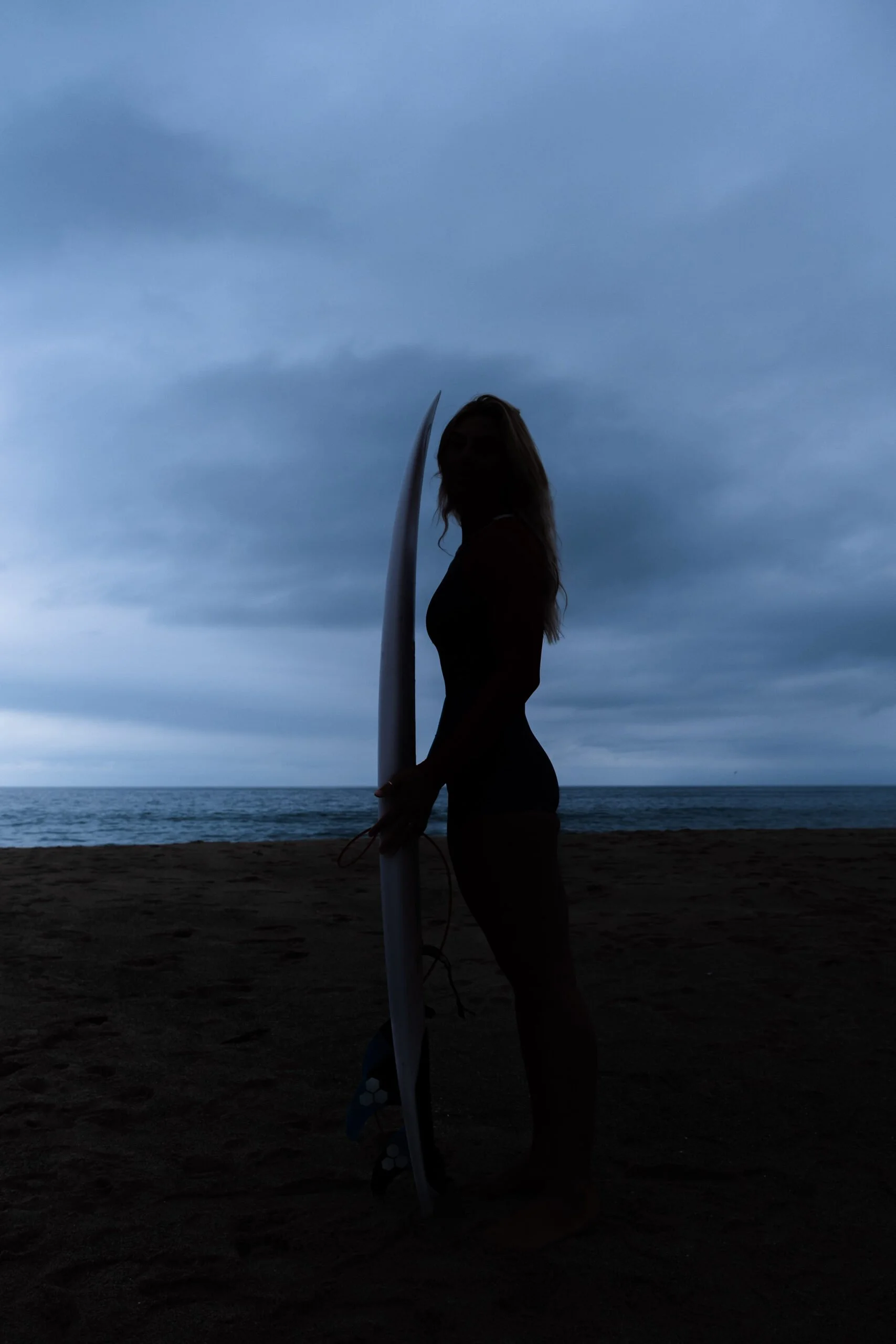 Silhouette of a woman holding a surfboard on the beach with ocean and cloudy sky in the background.