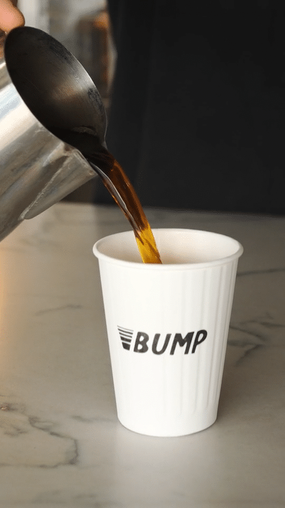 Hot coffee being poured from a metal carafe into a white paper cup with BUMP logo on a marble surface